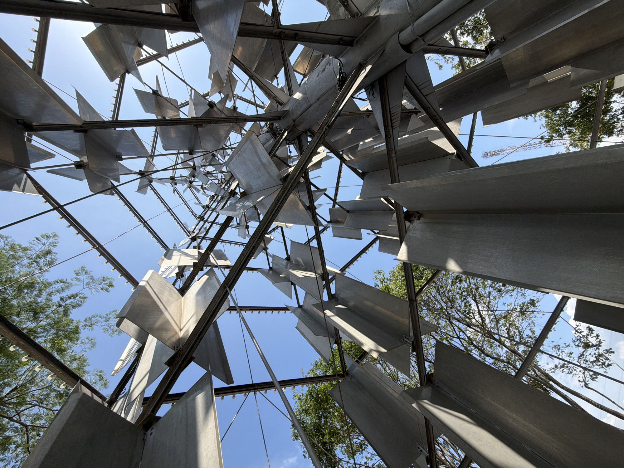 The image provides an upward-looking view through a large, intricate metallic structure, possibly a sculpture or architectural installation. The primary subject is composed of numerous silver-grey, flat, rectangular and trapezoidal panels, likely made of aluminum or steel, interconnected by a dark metal framework of beams and rods. This framework creates a complex, open geometric pattern that appears to taper upwards towards a central point.
Through the gaps in the metallic framework and panels, a clear, bright blue sky is visible, indicating a sunny day. Strong daylight illuminates the structure, casting distinct shadows and highlighting the angular forms and reflective surfaces of the metal. Lush green trees with visible branches and foliage are seen on the periphery and through some of the openings, suggesting an outdoor setting.
In one of the supporting crops, a construction crane is faintly visible in the distance against the sky, framed by the structure. No people are present, and no specific activity is taking place. The overall impression is one of modern design, geometric complexity, and a dynamic interplay of light and shadow. The context for this scene is Ciudad Guayana, Venezuela. There is no visible text in the image.