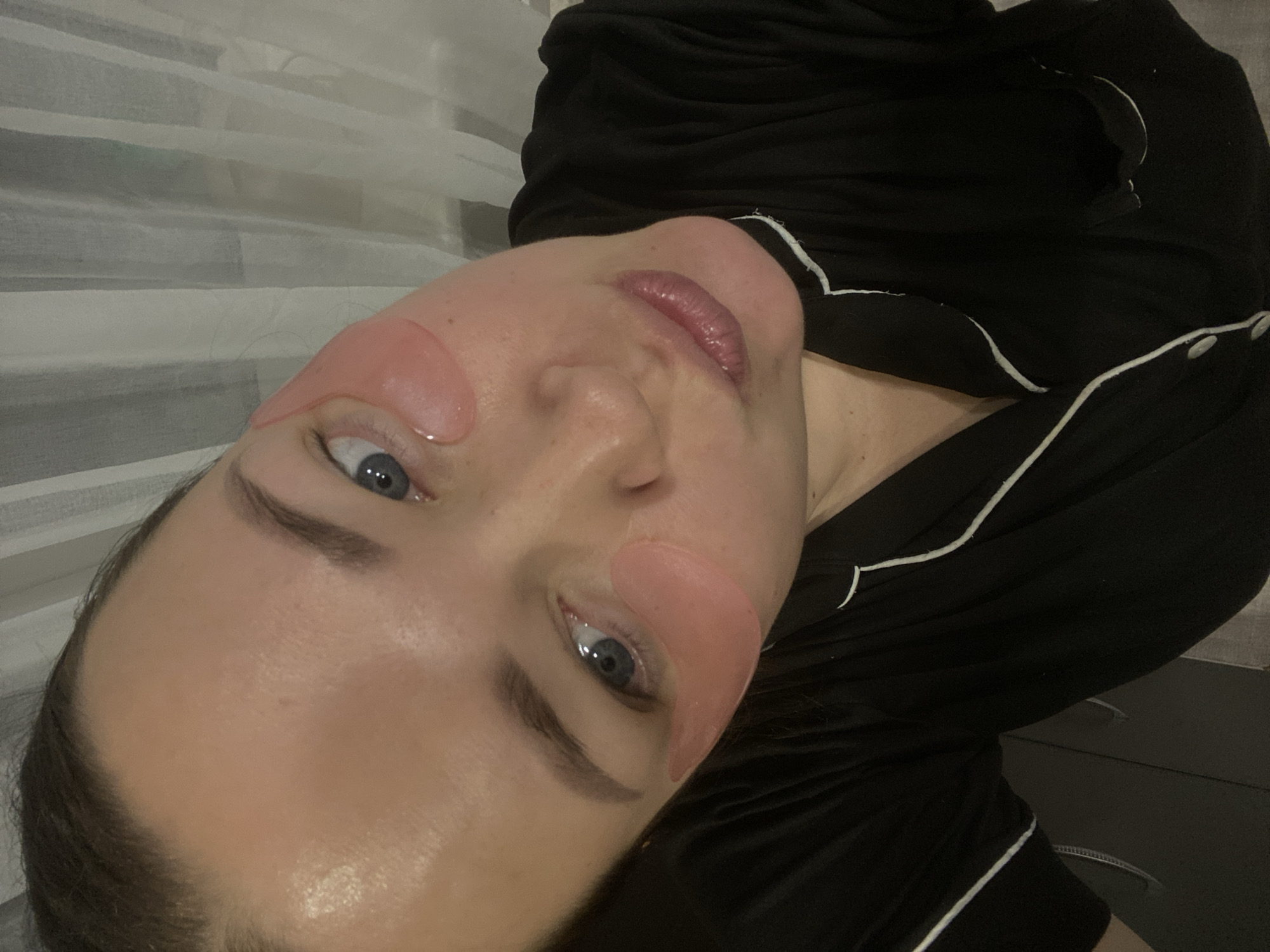 The image captures a close-up, high-angle portrait of an individual in what appears to be an indoor setting in Sochi, Russia. The subject is a fair-skinned person, likely female, with blue eyes, looking directly at the camera. Brown hair is visible on the upper left side of the frame. Two translucent pink gel under-eye patches are visible, one beneath each eye. The individual's lips are pursed, suggestive of a 'kissing' expression. They are wearing a black top, possibly pajamas, featuring white piping along the collar and front opening. The background consists of light-colored, vertically hanging fabric, consistent with curtains or drapes.