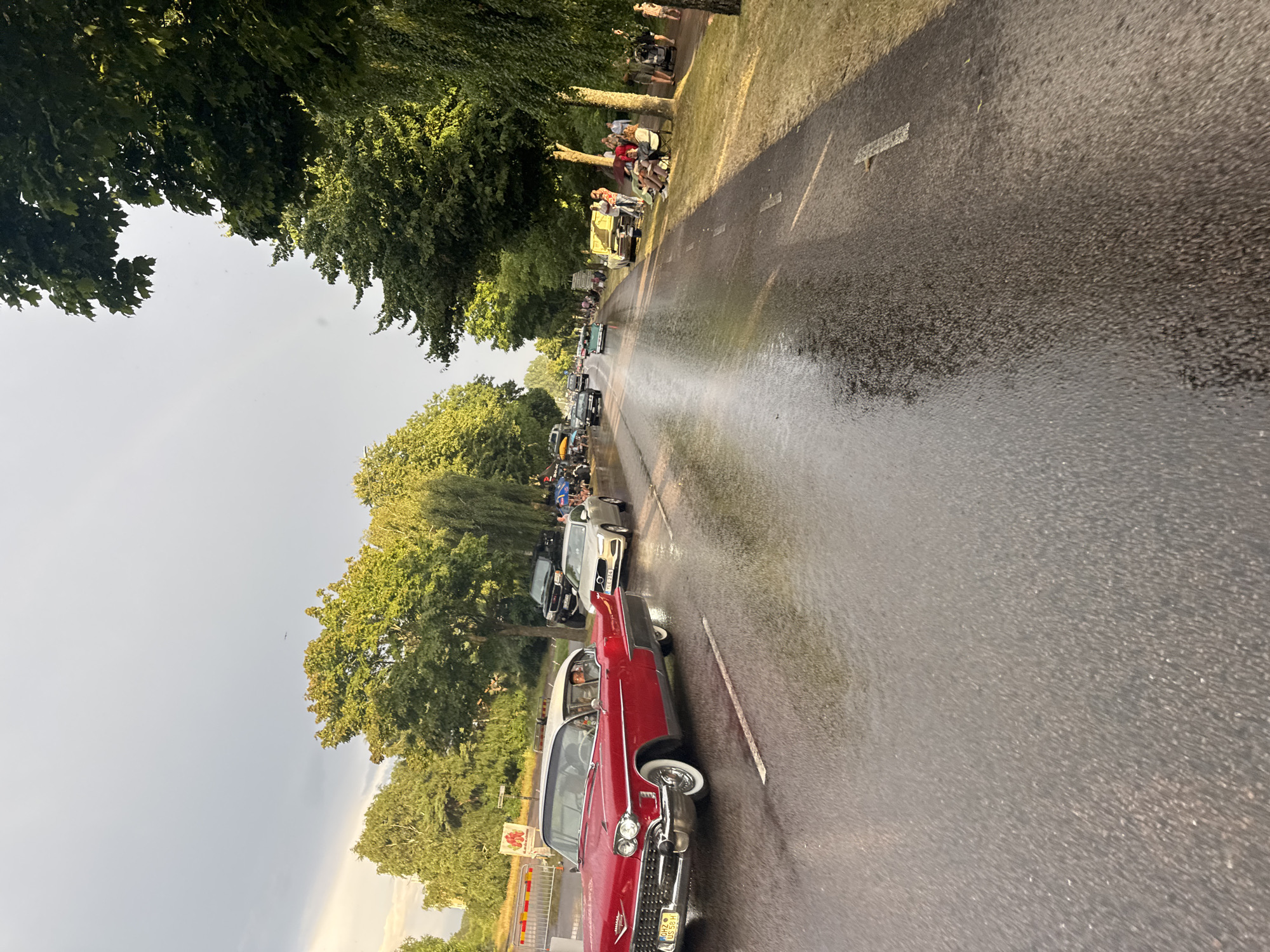 The image depicts a street scene in Falkenberg, Sweden, likely during a classic car event. A red vintage Cadillac is prominent in the foreground, driving on a wet asphalt road. Several other classic and modern cars are visible further down the road, along with a group of people gathered on the grassy verge to the right. The weather appears to be overcast, with trees lining both sides of the street. The presence of what looks like a barricade and signage suggests a designated event area. The road surface indicates recent rainfall, creating reflections from the sky and surrounding lights.
