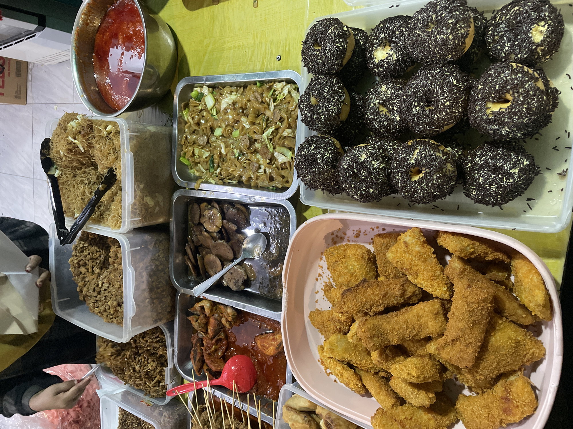 The image shows a street food stall in North Jakarta, Indonesia. The stall features a variety of Indonesian dishes, including mie goreng (fried noodles), perkedel kentang (potato fritters), and donuts with chocolate sprinkles. Other dishes include deep fried fish, deep fried tempeh, and fried eggplant. A pot of red sauce can be seen. A person can be seen preparing a food order.