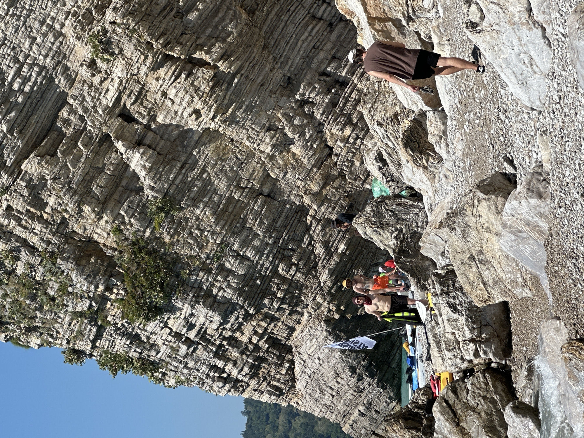 A group of people is near a large rock cliff near the water in Budva, Montenegro. Some people are standing near the water's edge, while one person is walking along the cliff face, higher up. A white flag is planted near the water. The sky is visible and blue. The water is not clearly visible.
