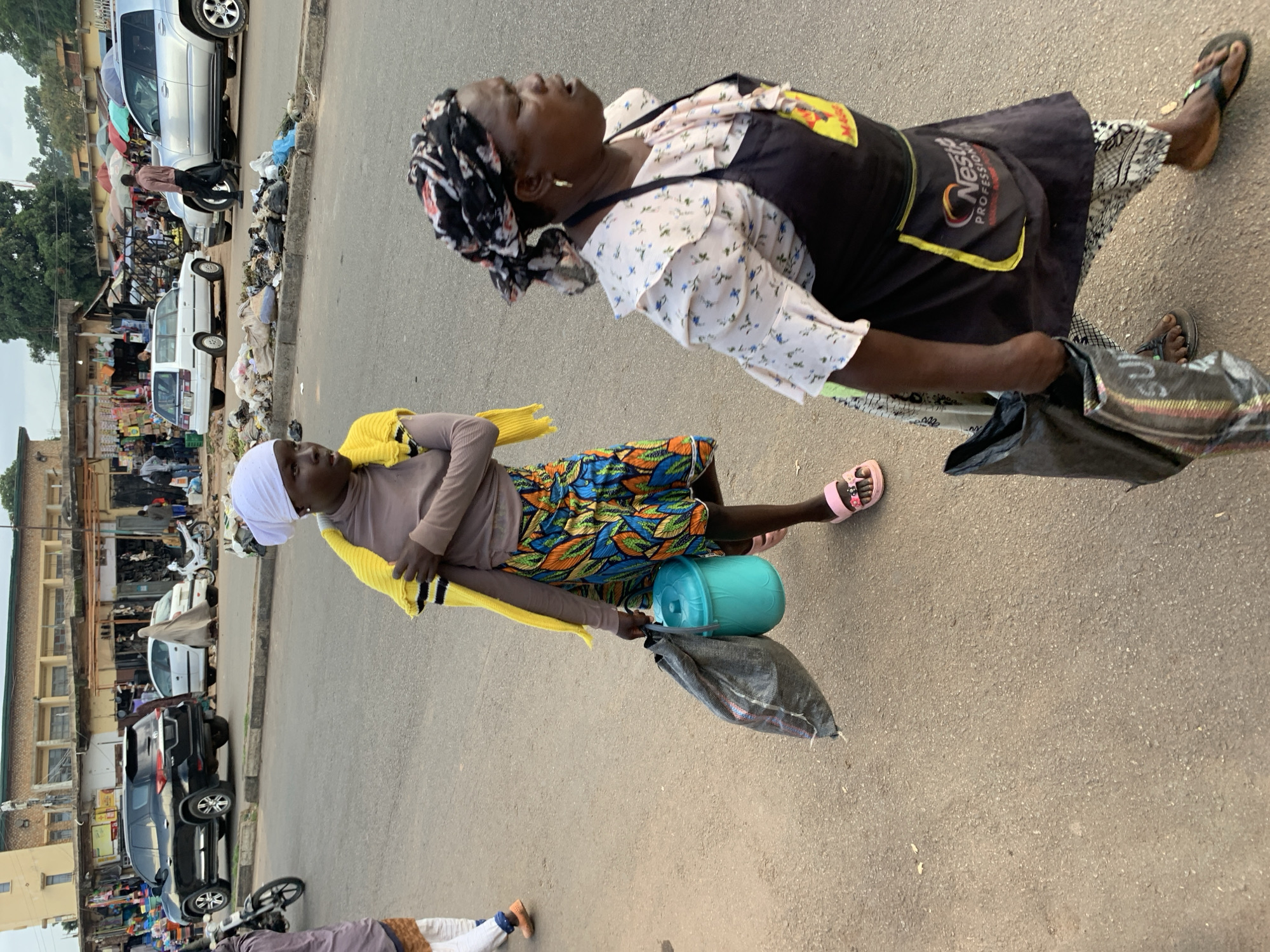 The image shows two women in what appears to be a busy street market in Jos, Nigeria. The woman in the foreground is wearing a light brown garment, a white headscarf, and a brightly colored patterned skirt. She is holding a yellow cloth and has a teal plastic container and a black bag at her feet. She is looking down. Behind her, another woman is visible, wearing a floral print blouse and a dark apron over a patterned skirt. She has a headscarf and is looking upwards. She is holding a black bag. The background shows market stalls, various vehicles including cars and motorcycles, and several people. The scene suggests a daytime setting with overcast conditions. The presence of market stalls and vehicles indicates a commercial area.
