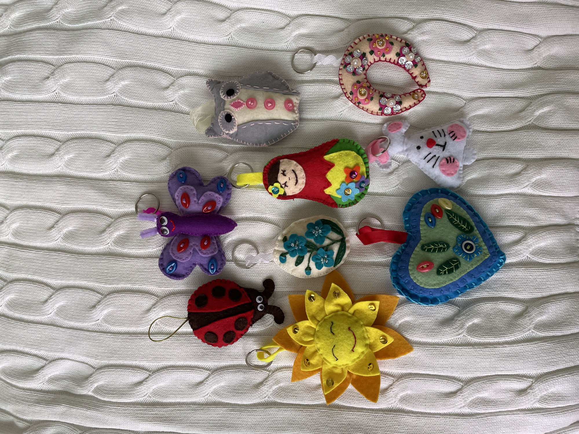 The image shows a collection of handcrafted felt keychains arranged on a white knitted fabric with a cable knit pattern. The keychains are various shapes and depict different themes, including an owl, a donut, a mouse, a butterfly, a matryoshka doll, an egg with flowers, a heart, a ladybug, and a sunflower. Each keychain has a metal keyring attached. The background is a textured white fabric, and the scene is captured in a well-lit, overhead shot. The city of Лесной Пейзаж Кп (шарапово, Russia is not depicted or implied in the visual information.