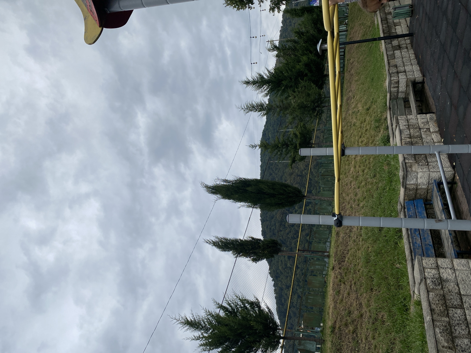 A street view captures an outdoor recreational area beneath an overcast sky in Lagodekhi, Georgia. The scene features a pull-up bar apparatus with yellow horizontal bars supported by grey vertical poles in the foreground. To the left, a weathered blue wooden bench is integrated into a low stone wall. Beyond this, a grassy incline rises, bordered by a green metal fence. A line of cypress trees stands along the fence line, with a lush, tree-covered mountainside in the background. Above, power lines are visible against the cloudy sky. On the far right, a portion of a structure with a yellow and red curved roof, possibly part of a playground or park feature, is visible. The ground is covered with dark, textured rubber matting, typical of a sports or play area. The overall atmosphere is subdued due to the overcast weather.
