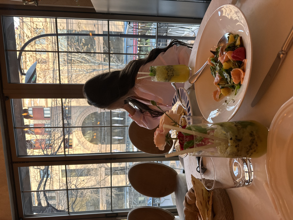 The image displays an indoor restaurant setting with a view of an urban street in Yerevan, Armenia. A female individual, seen from behind with long, dark, wavy hair, is seated at a white-linened table, looking out through large, gridded windows. She wears a light-colored, possibly pink, long-sleeved top, with her left hand near her face.
On the table in the foreground, a large white plate contains a salad composed of green leaves, orange segments, and red elements, with a silver spoon resting within. To the left, a tall glass holds a green and yellow beverage with visible kiwi seeds and mint, garnished with two white straws. An empty clear glass tumbler sits beside it. Further left, a woven basket contains assorted breads, including flatbreads and croissants. A small floral arrangement of light pink roses and foliage is presented in a vase, next to a blue and white patterned plate holding cheese slices. Another similar green and yellow drink is positioned further back on the table. A silver knife is placed to the right of the salad plate.
Through the windows, a multi-story building featuring reddish-brown (likely pink tuff stone) construction with large arched windows on its lower level is visible across the street. Bare deciduous trees and a curved black street lamp are also present in the exterior view. The architecture of the visible building is characteristic of central Yerevan, suggesting the location is near Republic Square or along a major thoroughfare. The scene is illuminated by warm, golden sunlight from the late afternoon or early evening.