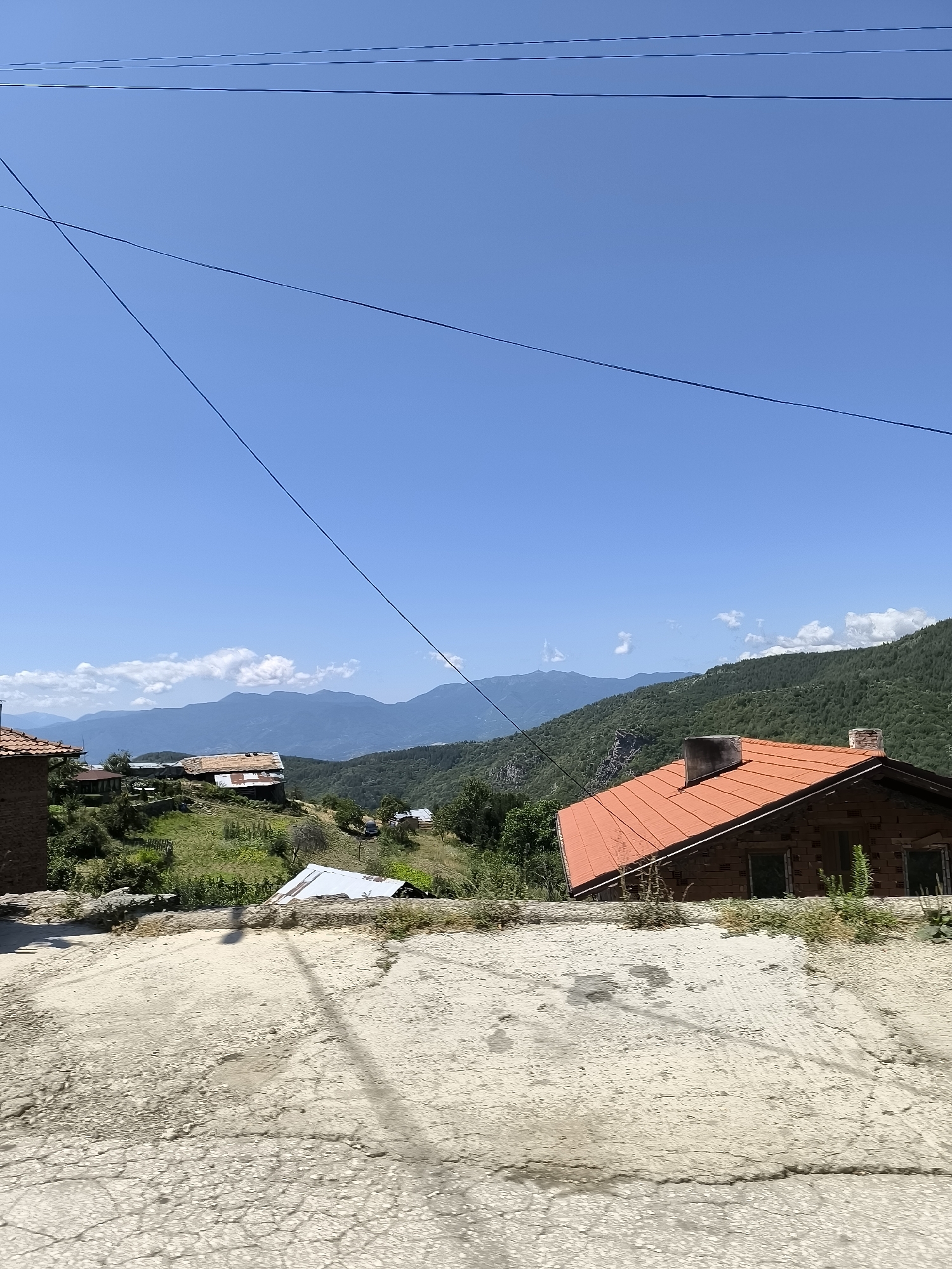 The image depicts a rural landscape in the village of Leshten, Bulgaria, under a clear blue sky with scattered white clouds. The foreground features a cracked and worn concrete surface, possibly a road or pathway, casting a faint shadow of overhead wires.
In the mid-ground, several rustic houses with red-tiled roofs are nestled on a hillside covered with green grass and scattered trees. One prominent house, built of stone and brick, has a distinctive chimney and a large orange roof. Further down the slope, other structures with weathered roofs are visible, suggesting a traditional village setting.
The background is dominated by rolling hills and distant mountains, covered in dense green forests. The overall scene conveys a sense of peace and tranquility, characteristic of a remote mountain village. No people or animals are visible in the image.