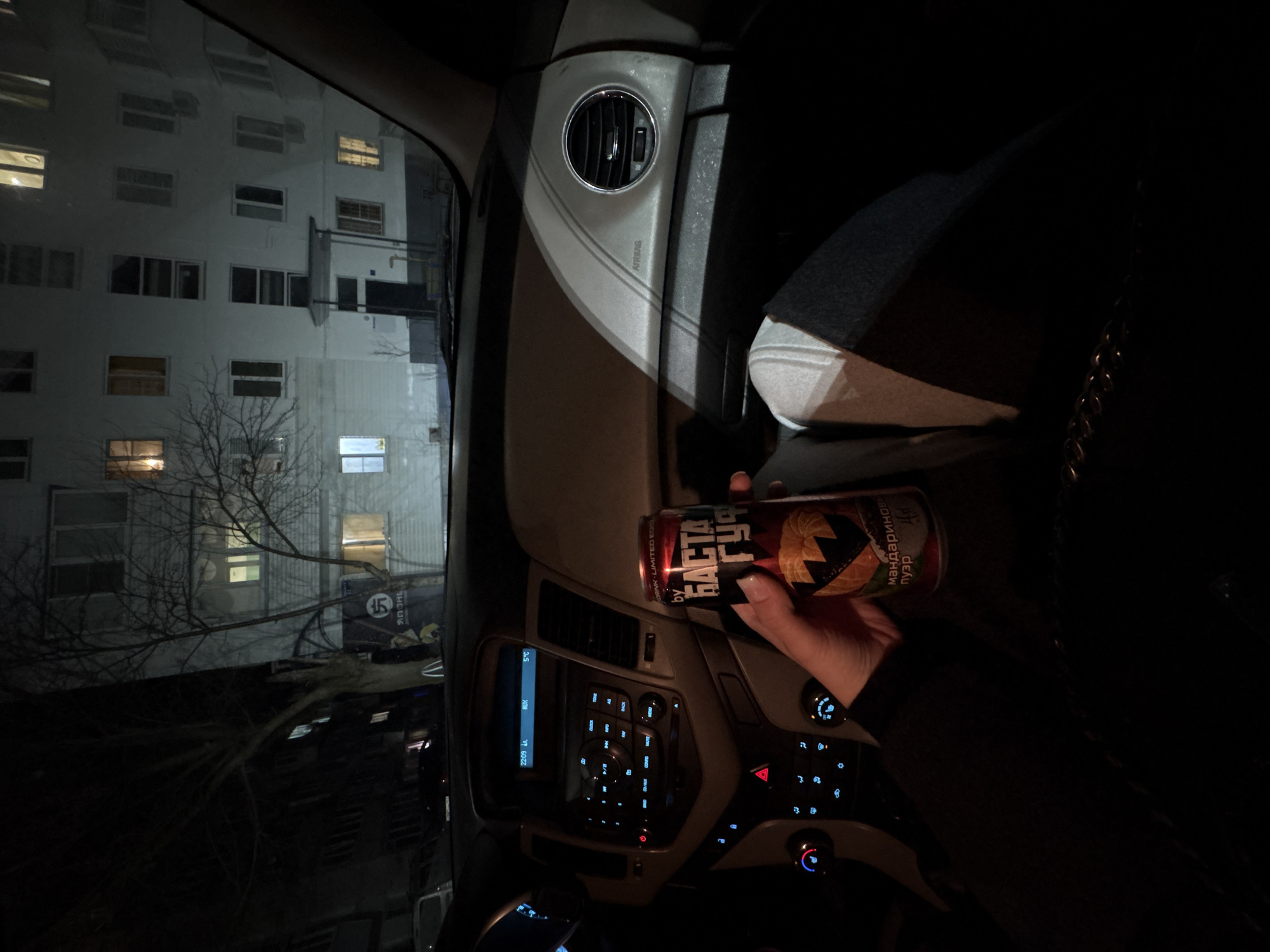 Here's a breakdown of the image:
**Location:** Appears to be inside a car, in what seems to be the city of Chaikovsky, Russia. This is inferred from the Cyrillic text on the drink can. A building is visible through the car's window.
**Subjects and Objects:**
* A person's hand is holding a can of a drink. The can has Cyrillic text, including the brand "БАСТА Гуд" and the phrase "мандариновый пуэр".
* The interior of a car is visible, including the dashboard, air conditioning vents, and a control panel with buttons.
* Outside the car window, there is a multi-story building with windows, some illuminated. Bare branches of trees are also visible.
**Environment:**
* It is nighttime. The lighting is dim inside the car, and the outside scene is illuminated by streetlights and lights from the building.
* The scene appears to be urban, as evidenced by the presence of the building.
**Action:**
* A person is holding a beverage inside a vehicle.