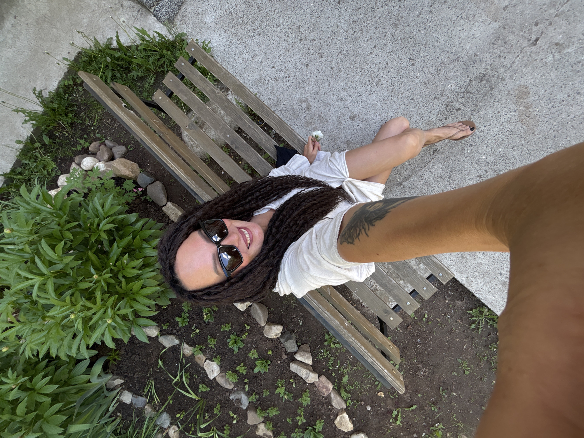 A medium-shot photograph shows a woman lying on a weathered wooden bench in what appears to be a park or garden. The angle is a slightly tilted overhead shot, possibly a selfie taken with an outstretched arm. The woman has long, dark hair styled in braids or locs, and she is wearing sunglasses. She is dressed in a light-colored, possibly white, dress or top and is holding a small white flower. Part of her tattooed arm is visible, closest to the camera.
The bench is surrounded by foliage, including a green leafy bush and areas of grass and small plants. Rocks are scattered around the bench's base, adding to the natural setting. The ground beneath the bench is a mix of soil and sparse vegetation. A paved area is adjacent to the bench, indicating a pathway or sidewalk.
The location is identified as Chaikovsky, Russia, based on source metadata. The overall impression is of a relaxed, outdoor scene.