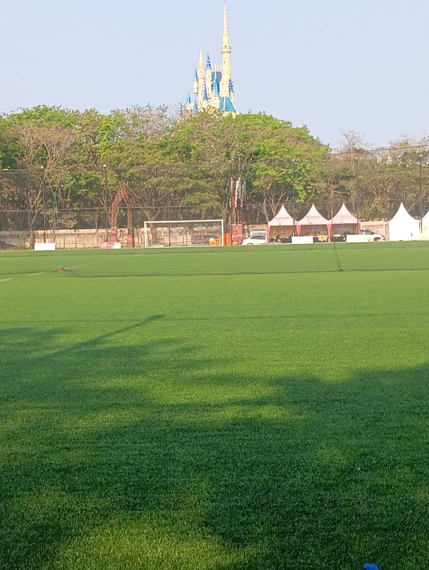 The image depicts a wide, artificial turf sports field, likely a soccer field, in Surabaya, Indonesia. In the background, behind a fence and a line of trees, is a distinctive castle-like structure with blue spires, resembling Cinderella Castle at Disneyland. Several white tents are set up in front of a low wall, with cars parked behind them. The sky is clear and light blue. The foreground is dominated by the green expanse of the field, with shadows from unseen objects cast across it. There are no people clearly visible in the image. The overall impression is of an open recreational space adjacent to a themed attraction.