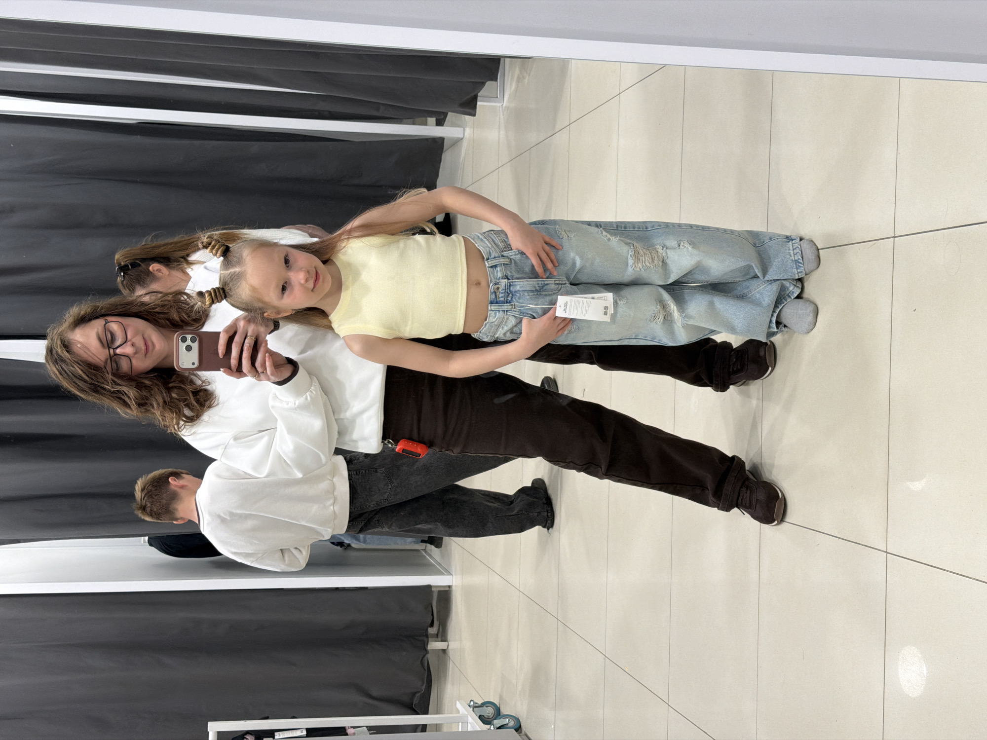 A woman with glasses is taking a photo of a child in a dressing room. The child is wearing a yellow top and ripped jeans. The dressing room has a mirror and a gray curtain. There are two other people in the dressing room, one partially visible. The floor is tiled. The photo is taken in a store in the city of Chaikovsky, Russia.