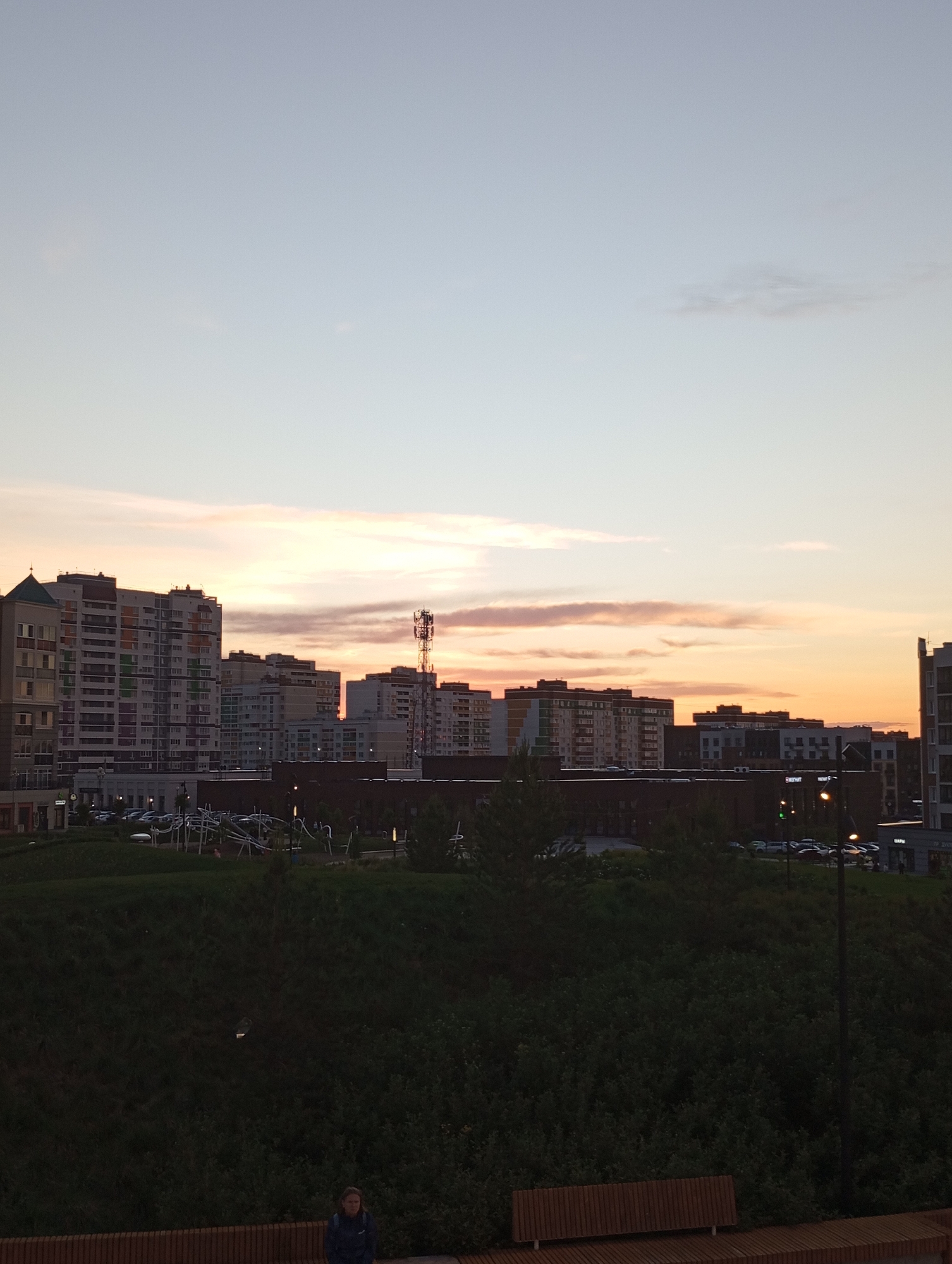 The image captures a skyline at sunset in what appears to be an urban setting. The scene includes several tall buildings with a variety of architectural styles, suggesting a modern or recently developed area. A telecommunications tower rises among the buildings.
Below the skyline, a park or green space is visible, with dense vegetation covering a raised area, possibly a hill or embankment. In the foreground, a wooden bench is positioned, and a person wearing a jacket stands beside it.
The sky is colored with the hues of sunset, blending from a light blue-gray at the top to orange and pink near the horizon. This color palette suggests the image was taken during either dawn or dusk.
Given the reference to Pervomaysky, Russia, it can be inferred that this image depicts an area within or near that city.