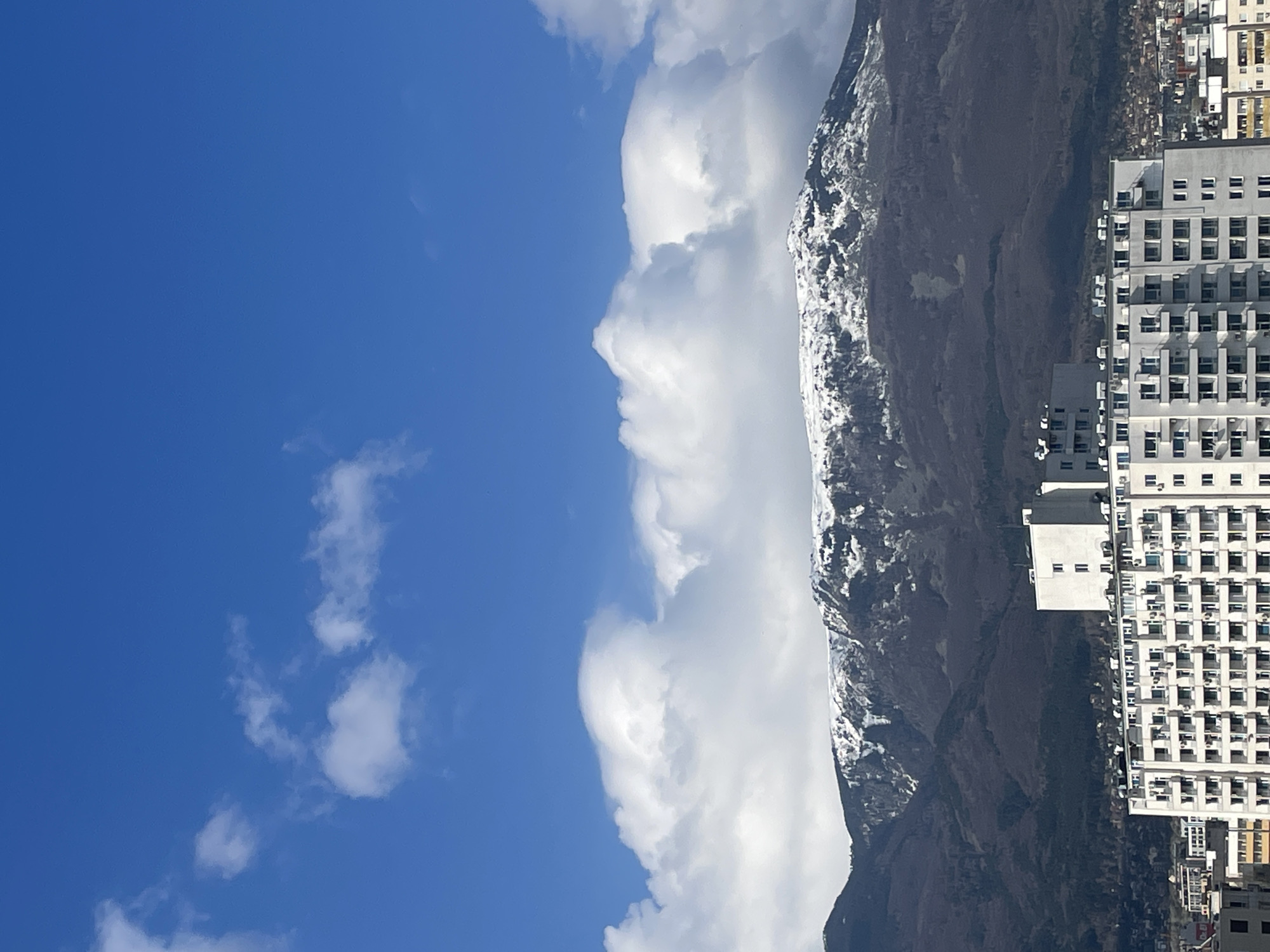 The image shows a mountain range with some snow on its peaks, next to buildings. There is a clear blue sky with white clouds. The location is most likely near Sofia, Bulgaria. The buildings appear to be residential and commercial, with many windows. The mountain is primarily rocky and barren, with patches of snow at higher elevations. The buildings seem to be situated near the foot of the mountain.
