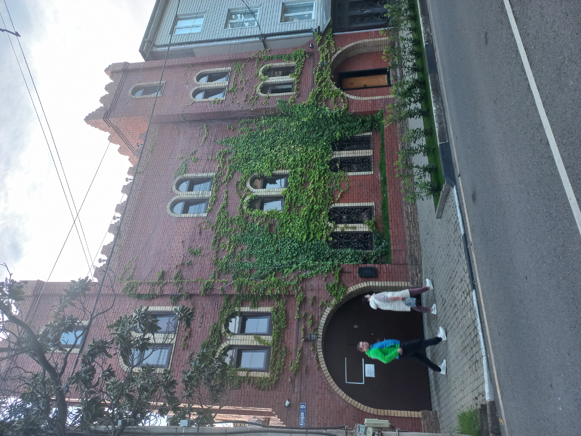 The image shows a red brick building covered in ivy, with unique arched windows. The building appears to be located in the city of Sochi, Russia, as suggested by the street sign on the building, which displays Cyrillic text. Two people are walking along the sidewalk next to the building. The building is of a unique architectural style, with what appears to be decorative elements. A paved sidewalk runs adjacent to the building, and a street is visible to the right.