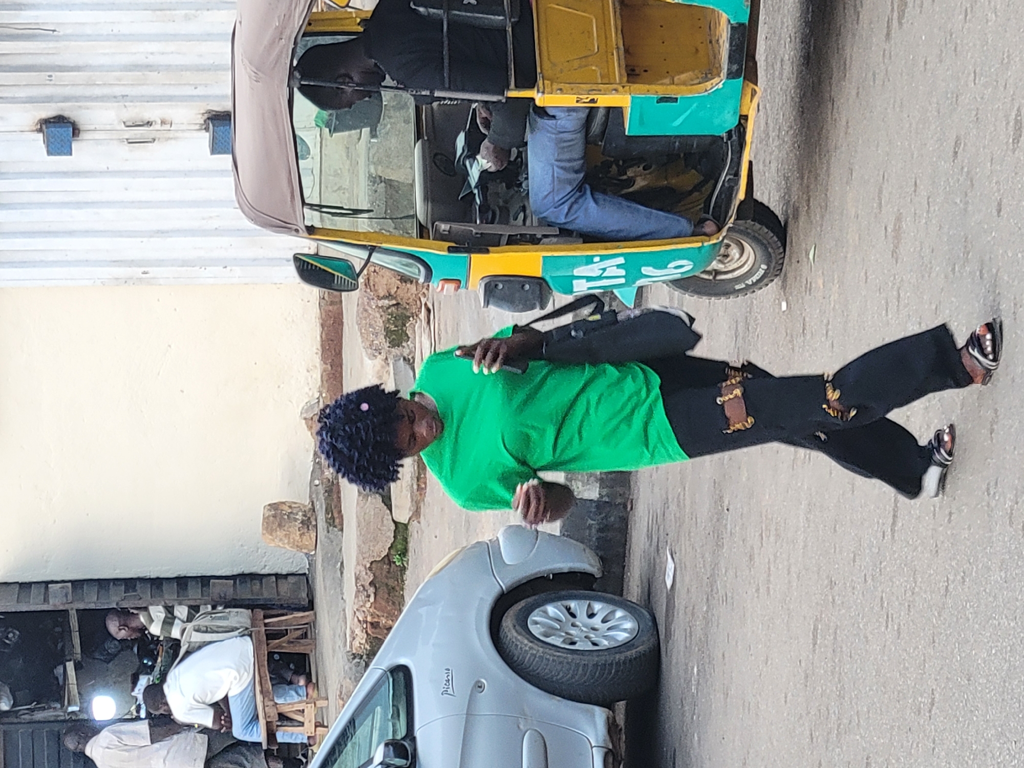 This is a slightly low-angle, full-shot photograph of a street scene in Barakin Lerawi, Nigeria. The image is oriented vertically. In the foreground, a silver car, possibly a Citroën Picasso, is parked on the right side of the frame. Its front fender and wheel are visible. To the left of the car, a man in a white shirt and blue jeans is bending over. Behind him, another man in a striped shirt is also bent over, and a third person in a white garment is seated.
Further back, a woman with dark curly hair, wearing a bright green t-shirt and black pants with a pattern of brown figures, stands and appears to be walking or preparing to move. She is holding a dark bag. Behind her, a yellow and green auto-rickshaw is parked. A man in a black shirt and blue jeans is seated in the driver's position of the auto-rickshaw. The auto-rickshaw has the number "41" written on its side in white.
The scene appears to be taking place during the daytime, under what seems to be clear weather. The ground is paved, suggesting a street or road. There are buildings and some structures visible in the background, indicating an urban or semi-urban setting. The overall impression is of a busy street with people going about their daily activities.