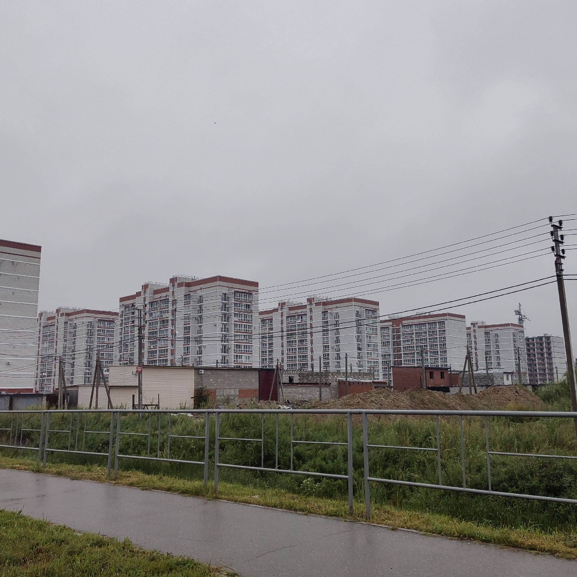 The image depicts a cloudy, overcast day in Chigiri, Russia, with a cluster of modern, multi-story apartment buildings dominating the background. The buildings are light gray with red accents around the upper floors and windows, and appear to be under construction or recently completed, as indicated by a construction crane visible in the distance. In the foreground, a paved walkway or road runs parallel to a metal fence, beyond which is a strip of overgrown grass and vegetation. Several utility poles and power lines are strung across the scene, also in the background. The overall impression is of a developing urban or suburban area.