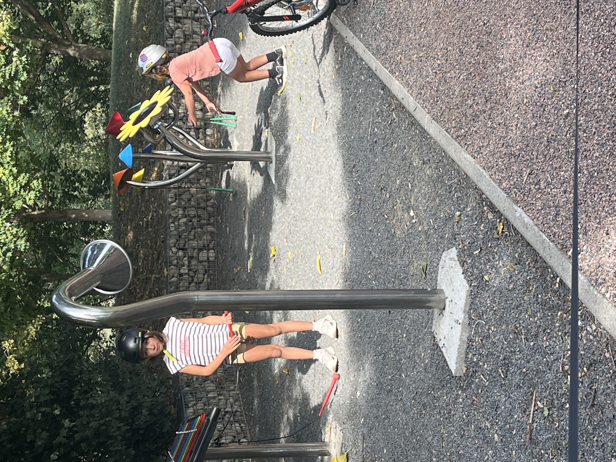 In Andrézieux-Bouthéon, France, two children are observed in an outdoor recreational area. The child on the left, wearing a striped t-shirt and shorts, stands near a musical instrument resembling a xylophone. They are also holding a red mallet. The child on the right, dressed in a pink t-shirt and white shorts, is interacting with a colorful, flower-shaped structure, also possibly a musical instrument, and is holding jump ropes. A red bicycle is parked to the right of the frame. The ground is covered with gravel, and the background features trees and a stone wall.