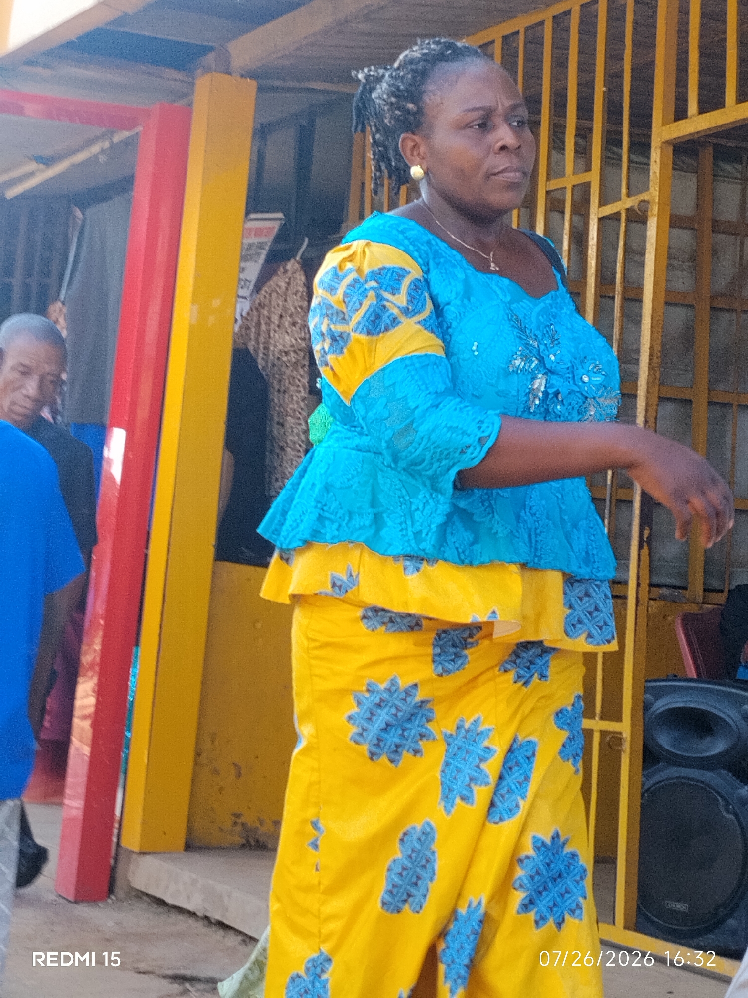 In Umuode, Nigeria, a woman stands outdoors in front of a yellow metal gate. She is wearing a vibrant blue and yellow patterned dress. Her hair is styled in braids, and she has on a gold earring. To her left, a portion of a blue t-shirt is visible on another person. Red and yellow poles are also present in the foreground, along with a black speaker. The ground appears to be concrete and dirt. A timestamp in the bottom right corner reads "07/26/2026 16:32". The text "REDMI 15" is visible in the bottom left corner.