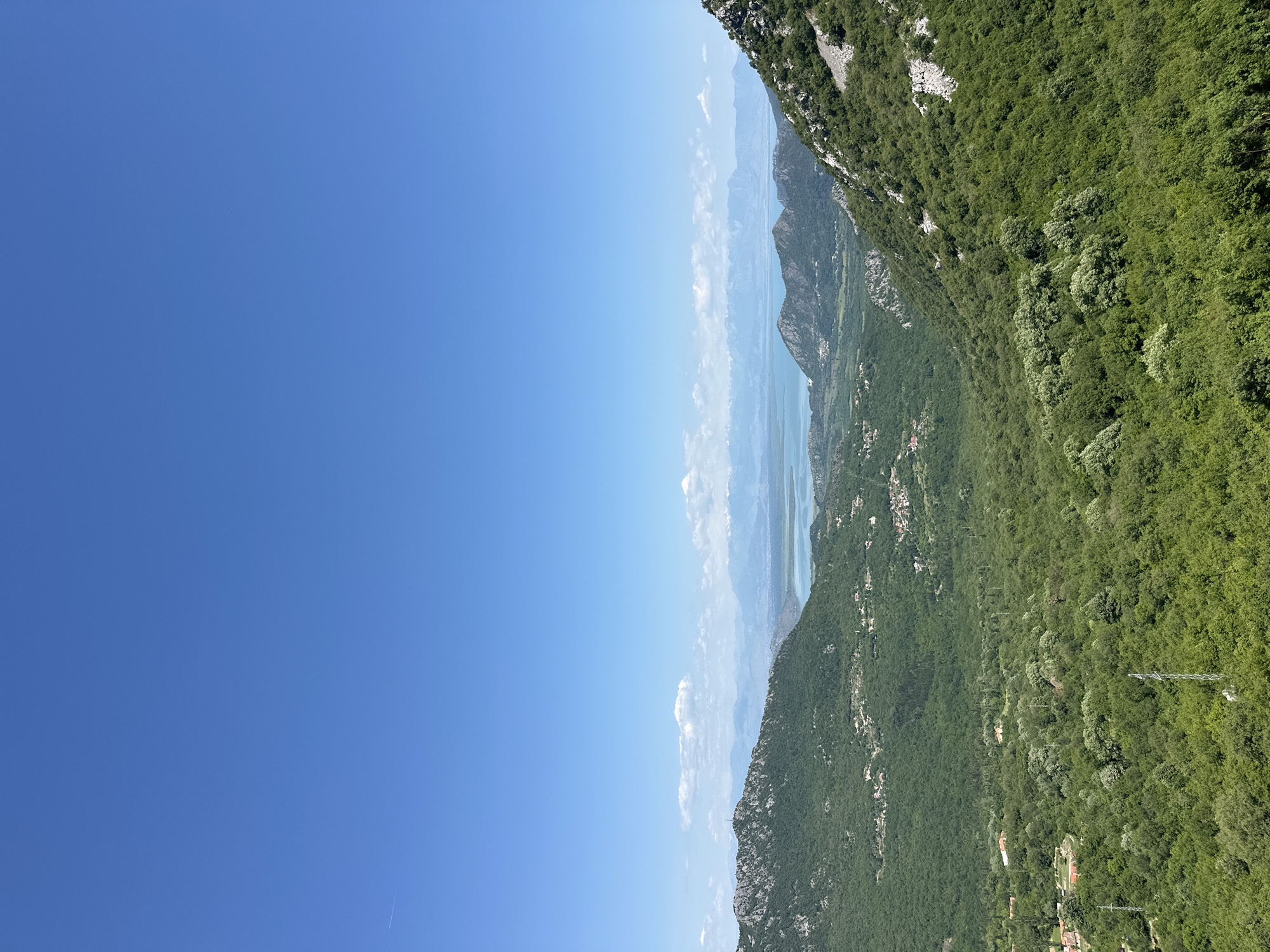 The image depicts a scenic view from Bukovik, Montenegro, showcasing a mountainous landscape under a clear blue sky. The terrain features lush green vegetation covering the hillsides and slopes, giving way to rocky formations and cliffs. In the distance, a body of water can be seen nestled between the mountains, partially obscured by haze or clouds. A few structures, likely buildings, are scattered across the lower slopes. Power lines are visible amidst the vegetation.