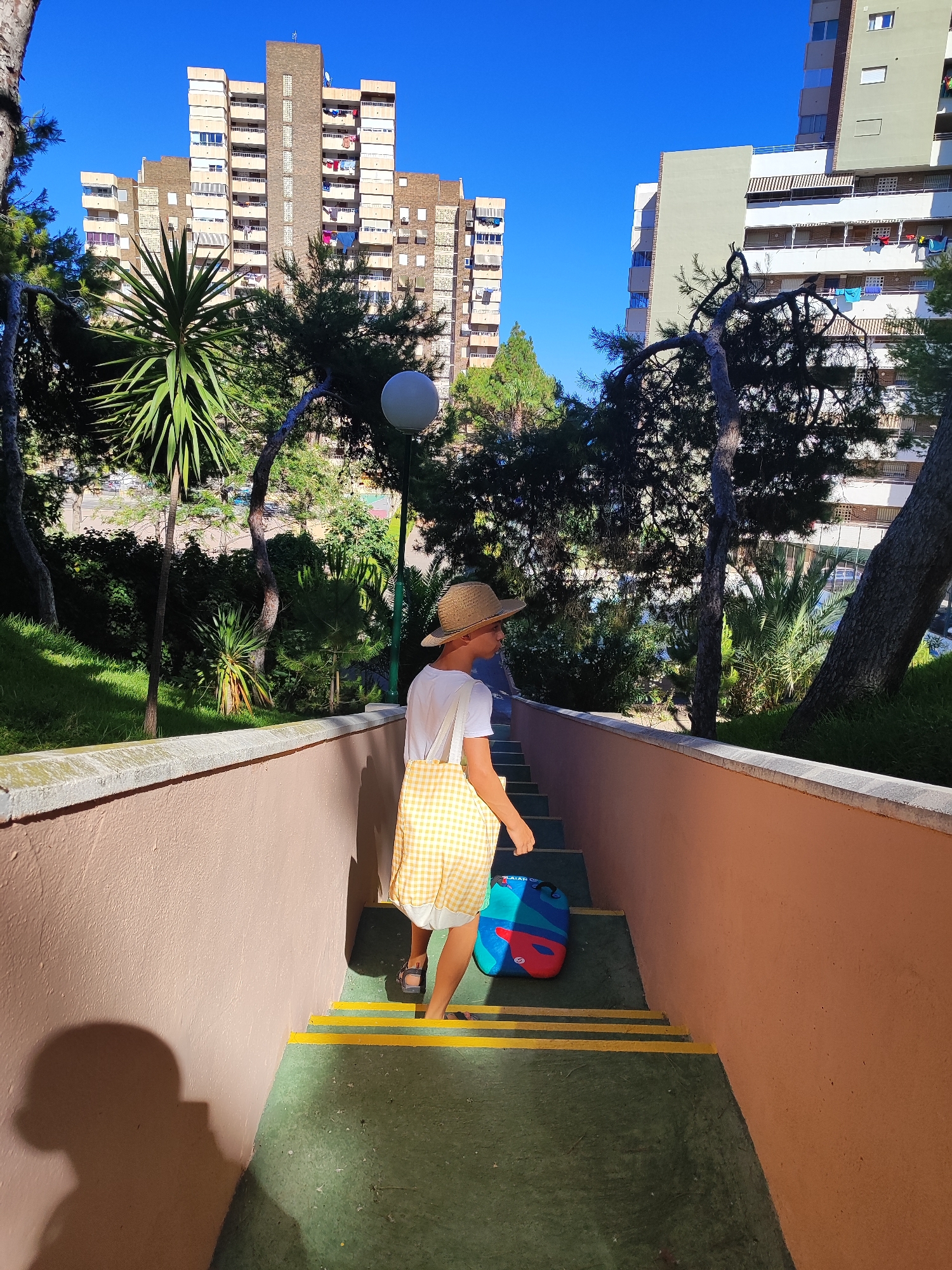 A child wearing a straw hat, white t-shirt, and yellow checkered shorts walks down an outdoor staircase. The child is carrying a colorful bag. The staircase has green steps with yellow safety strips. The staircase is flanked by an orange-pink wall on the left and a lighter orange-pink wall on the right. Lush green vegetation, including palm trees and other foliage, surrounds the staircase. In the background, tall apartment buildings with visible balconies and laundry hanging out are seen against a clear blue sky. The scene appears to be set in a residential area, likely in a Mediterranean climate, consistent with the city of El Campello, Spain. The lighting suggests it is daytime, with sunlight casting shadows on the wall and steps.