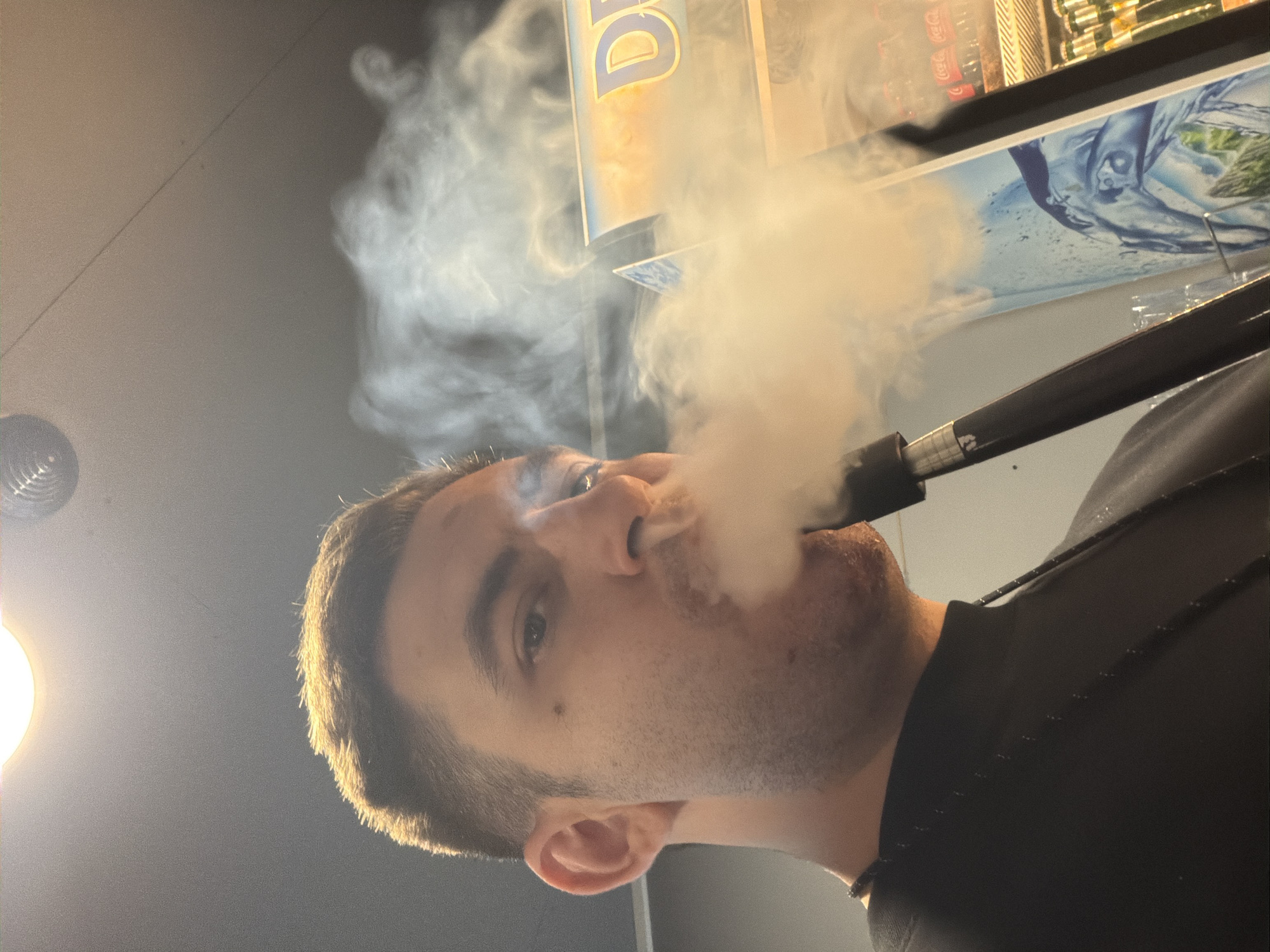 A man exhales a large cloud of white smoke, apparently from a vaping device or hookah. The location appears to be a store or cafe, based on the shelving with various drinks and a Coca-Cola advertisement visible in the background. The presence of the Greek letter "D" suggests this might be in or around Varna, Bulgaria, given the prevalence of that lettering in shop signs there. The man is wearing a black shirt, and the lighting appears to be artificial, originating from above and to the left. He is fair-skinned with short brown hair.