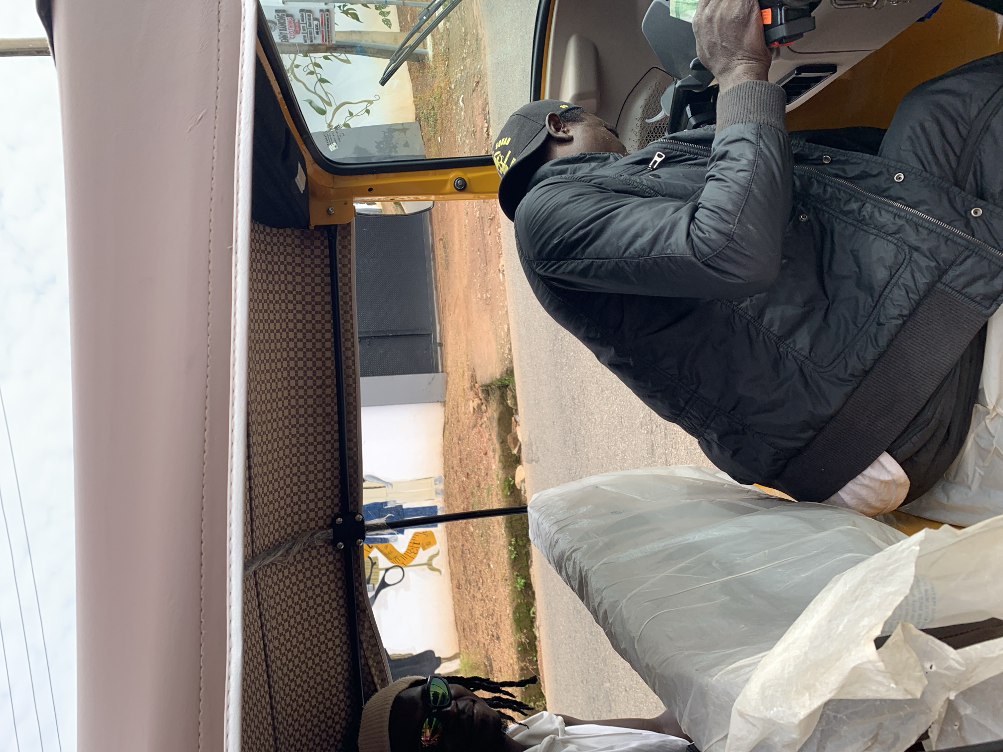 The image depicts the interior of a yellow auto-rickshaw, likely in the city of Gura Top, Nigeria. Two individuals are visible inside. In the foreground to the right, a man wearing a black jacket and a black baseball cap is seated in the driver's position, facing forward and holding the steering wheel with his right hand. His left arm is also visible. To the left of the driver, another person is partially visible, wearing sunglasses and a light-colored beanie. This person is looking towards the camera. The interior of the auto-rickshaw features a checkered pattern on the ceiling lining and a white plastic covering on the passenger seat. Through the open side of the vehicle, a dusty road and a building with a mural depicting sewing-related items can be seen. The overall scene suggests a candid moment captured during a journey.