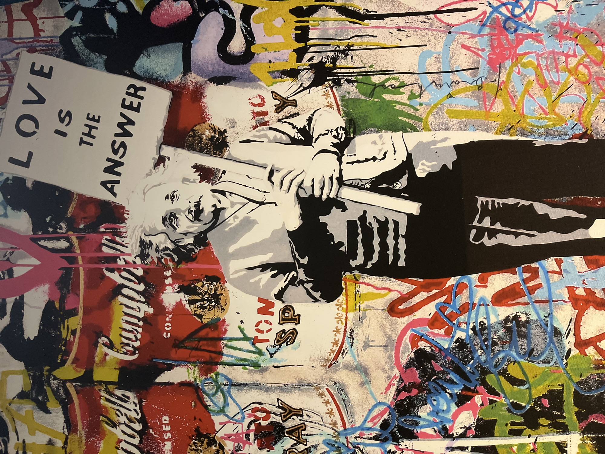 The image is a vibrant, mixed-media artwork. Prominently featured is a black-and-white portrait of a person, likely Albert Einstein, holding a sign that reads "LOVE IS THE ANSWER." This portrait is layered on top of a background featuring graffiti-style artwork, including elements such as Campbell's soup cans and other colorful abstract designs. The overall aesthetic is reminiscent of street art or pop art. The location, Grimaud, France, cannot be confirmed or denied based on the image. No specific clues within the image provide clear evidence of that location.
