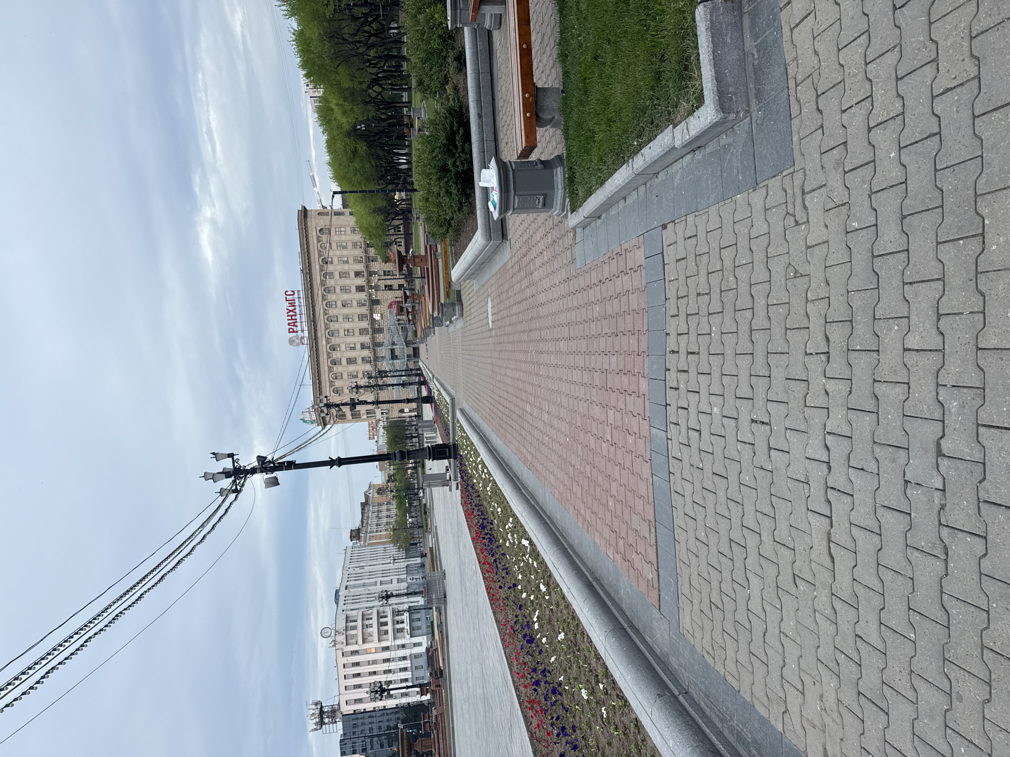 The image shows a paved walkway in Khabarovsk, Russia. The walkway is made of interlocking concrete pavers in two colors, gray and reddish-brown. The walkway is bordered on one side by a flowerbed with red, white, and purple flowers and on the other side by a line of benches and grass. In the background, there are buildings, including one with the Cyrillic text "РАНХиГС". Above the flowerbed is a light gray concrete edge, with a few segments of dark gray concrete separating the flowerbed from the walkway. Overhead, utility poles and wires stretch along the street. The sky is overcast.