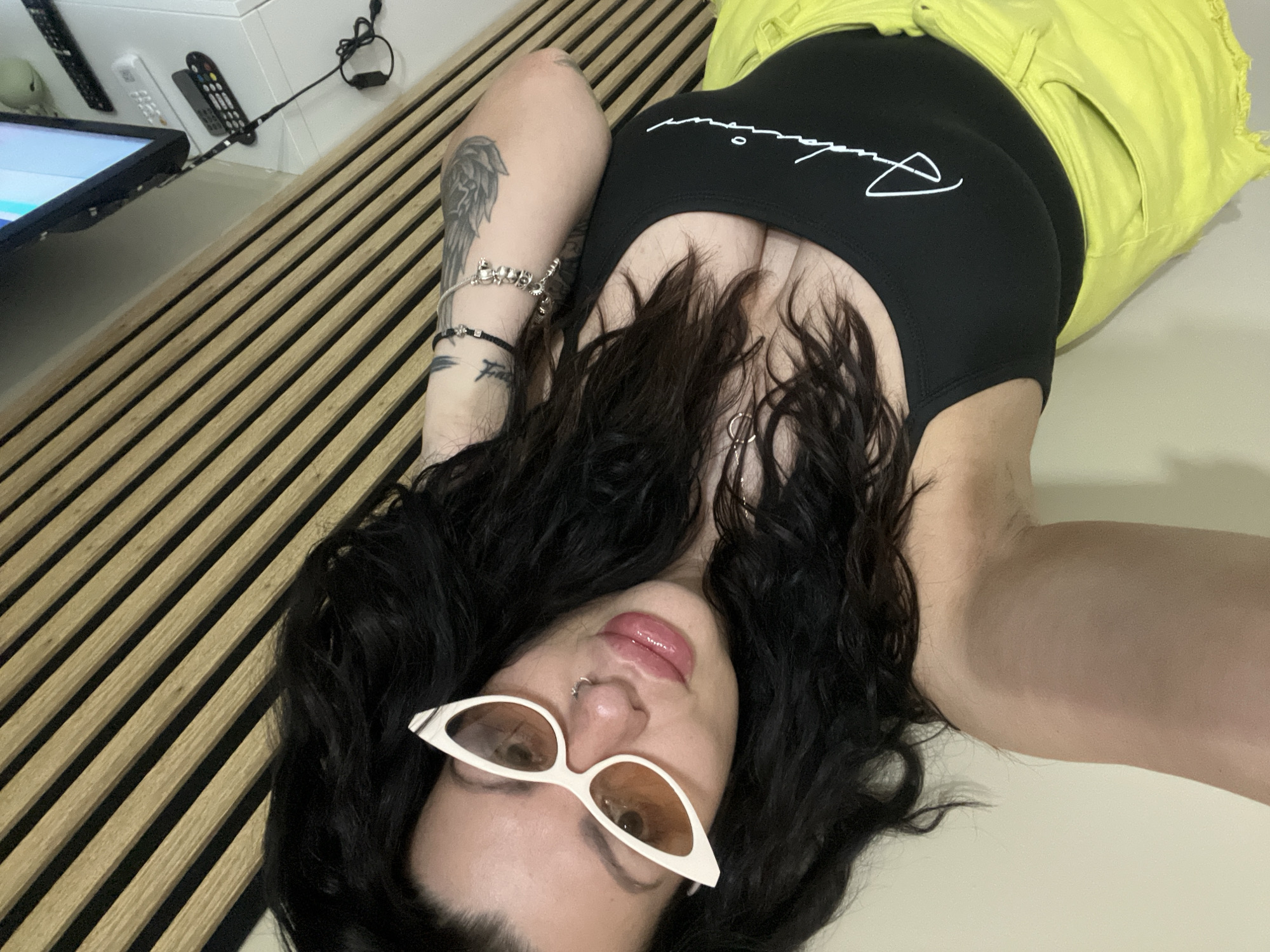 A woman lies on a bed or floor, facing the camera, with a wooden slat headboard behind her left arm. She is wearing a black top with white script and bright yellow shorts. She has dark curly hair, sunglasses with white frames and amber lenses, and jewelry on her left arm. A television and several remote controls are visible in the upper left corner. There is no evidence of the location being in the city of Plovdiv, Bulgaria.