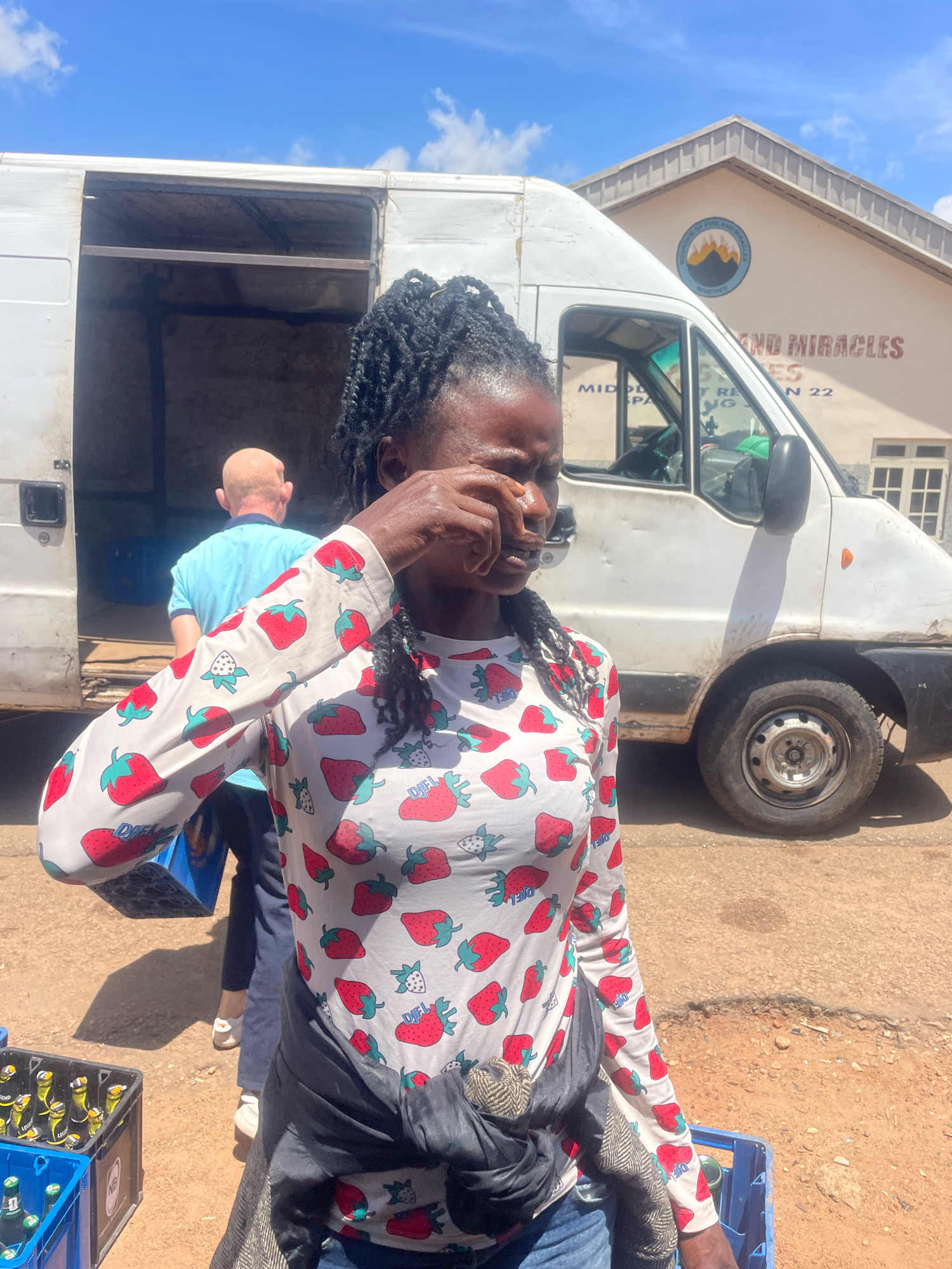 In Jos, Nigeria, a young woman stands in front of a white van, her face obscured as she wipes her eye. She wears a long-sleeved shirt adorned with a pattern of strawberries. In the background, a building with "MOUNTAIN OF FIRE AND MIRACLES MINISTRIES" signage is visible. The scene appears to be outdoors, with a dusty ground and crates of beverages in the foreground. The woman's expression suggests discomfort or distress.