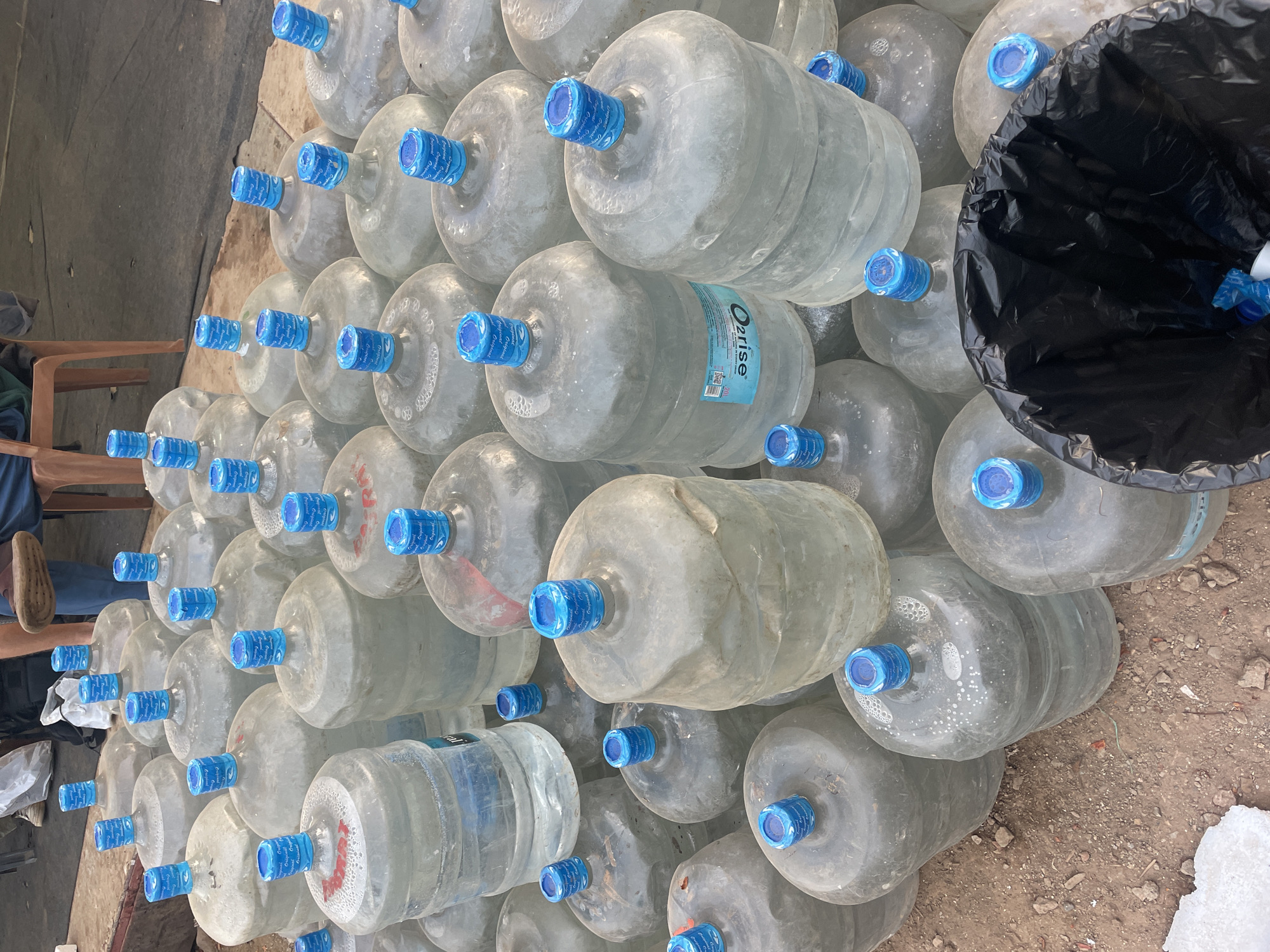 The image depicts a large stack of approximately 50 empty, transparent, reusable water bottles with blue caps. The bottles, possibly 5-gallon size, are arranged in a haphazard, multi-layered pyramid on the ground, with some bottles bearing a label reading "Ozrisse." A black trash bag is partially visible on the right side of the stack, seemingly containing some of the discarded bottle caps. The bottles are resting on an uneven, dirt or gravel-covered surface, possibly outdoors. Parts of a human being in a chair are visible in the frame. The scene is presumed to be in Mira-Bhayander, India.