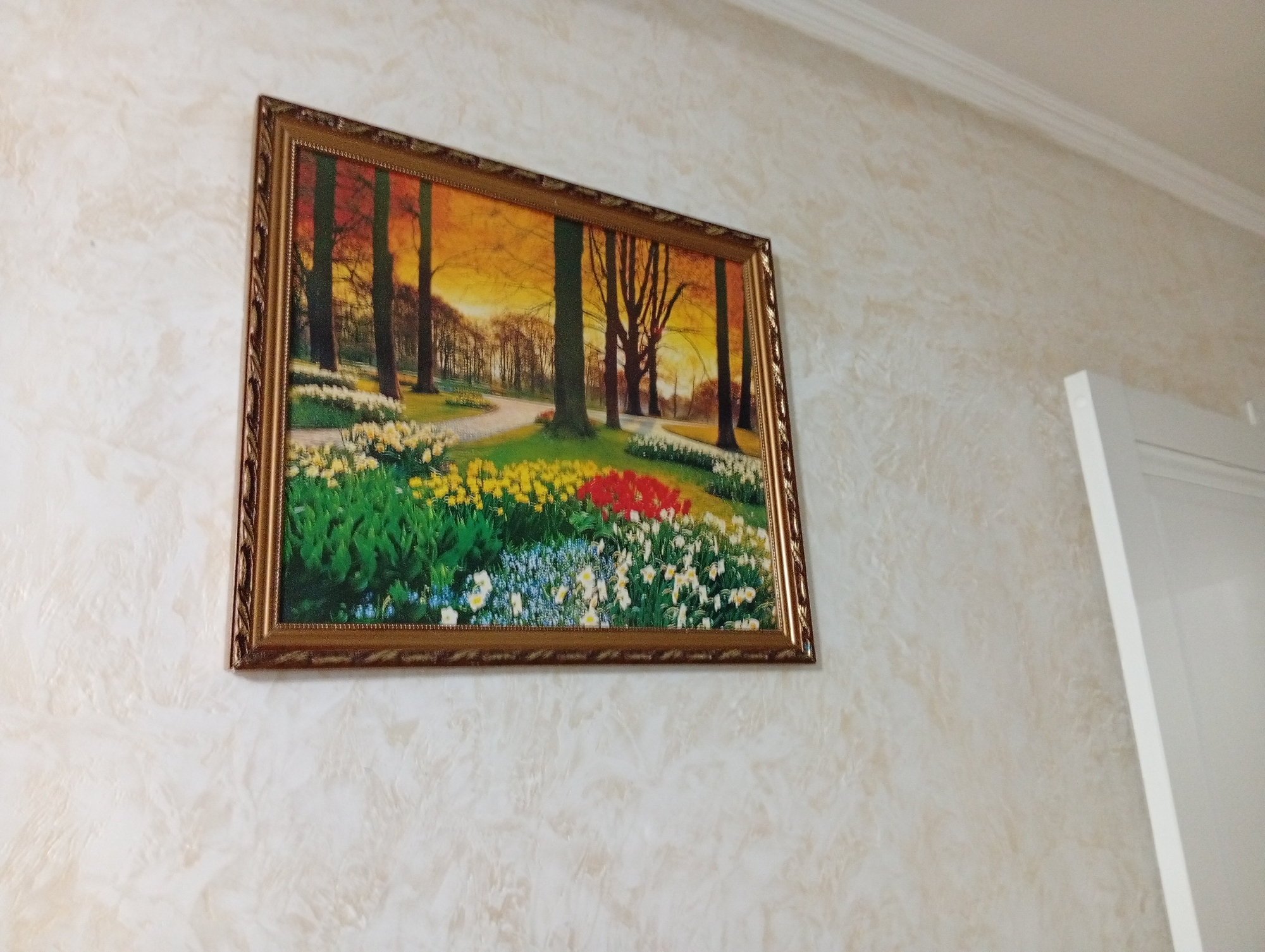 The image shows a framed painting of a park with trees and flowers, hanging on a textured beige wall. The painting depicts a path winding through a garden with a variety of colorful flowers, including red, yellow, and white. The background features trees with a golden sunset sky. To the right of the painting, a portion of a white door frame is visible. The overall setting appears to be an indoor environment, possibly a home or a public space. The location of Esik, Kazakhstan is not discernible from the image content.