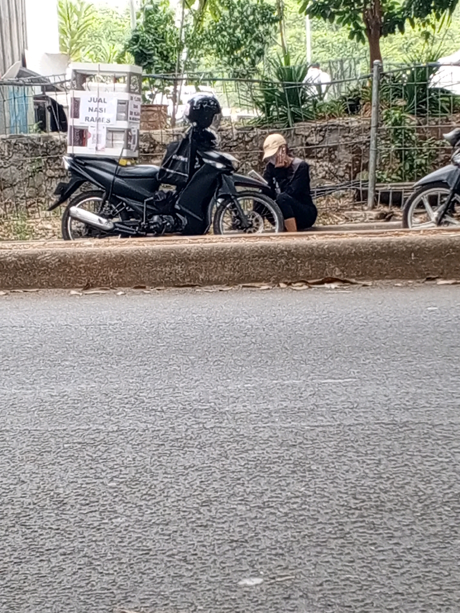 The image depicts a street scene in South Tangerang, Indonesia, with a black motorcycle parked on the side of the road. A person wearing a beige cap and dark clothing is crouched next to the motorcycle, holding their face with their hand. A white display case with text that appears to be "JUAL NASI RAMES" is attached to the back of the motorcycle. In the background, a stone wall and vegetation are visible, along with a fence. Another motorcycle is partially visible on the right side of the frame. The road in the foreground is asphalt.