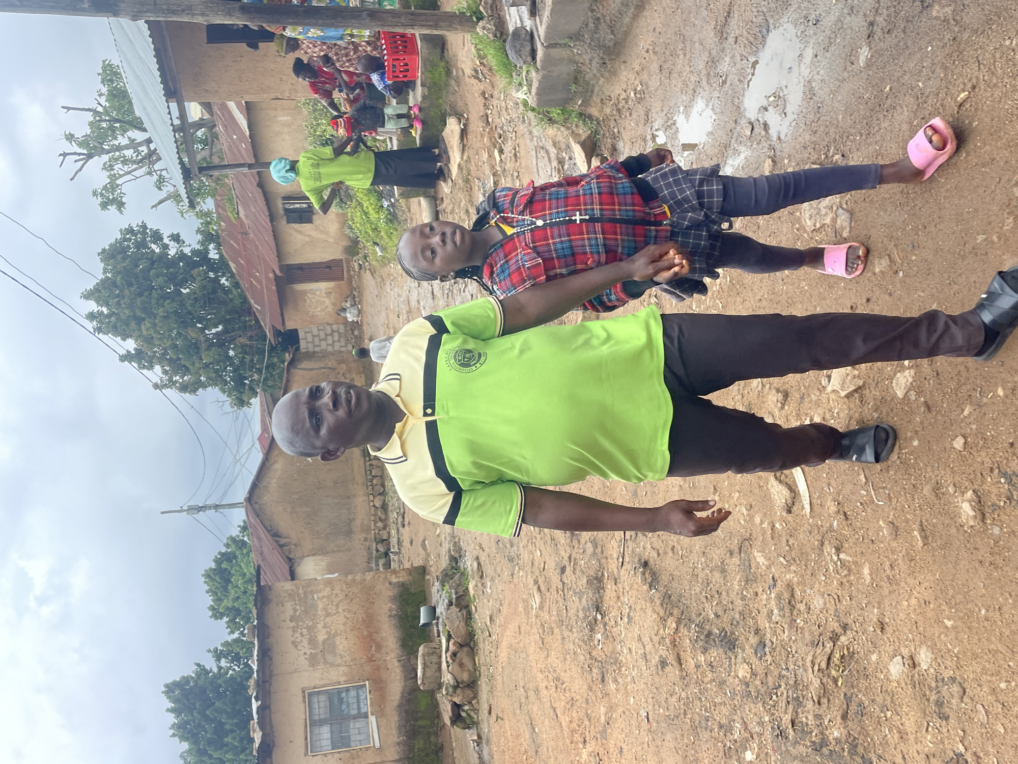 In Barakin Lerawi, Nigeria, an adult male in a lime green polo shirt with black and yellow trim and dark pants, wearing black sandals, walks on a dirt path holding the hand of a young girl. The girl wears a red, black, and blue plaid jacket over a yellow shirt, a dark pleated skirt, dark leggings, and pink slides. A white cross necklace is visible on the girl. In the background, several mud-brick buildings with corrugated metal roofs are visible, along with trees and an overcast sky. Other people, including a woman in a green headscarf and shirt and individuals in traditional attire, are present near one of the buildings.