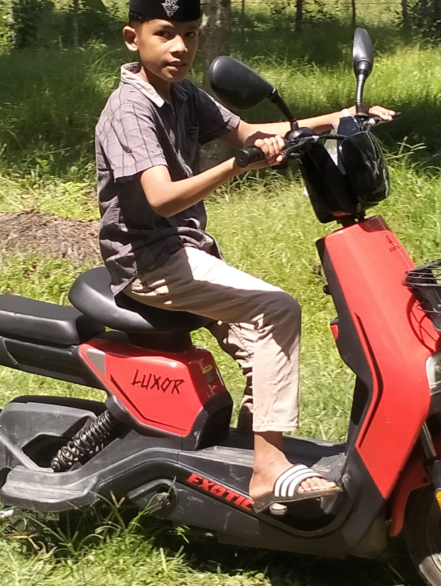 A young Indonesian boy with dark hair and wearing a black cap and a plaid shirt is sitting on a red and black electric scooter in Meunasahalue, Indonesia. He is holding the handlebars, and his feet are resting on the scooter's footboard. The scooter has the word "LUXOR" written in red on its side. The boy is wearing beige pants and white and black striped sandals. The background consists of lush green grass and trees. The lighting suggests it is daytime, with sunlight casting shadows.