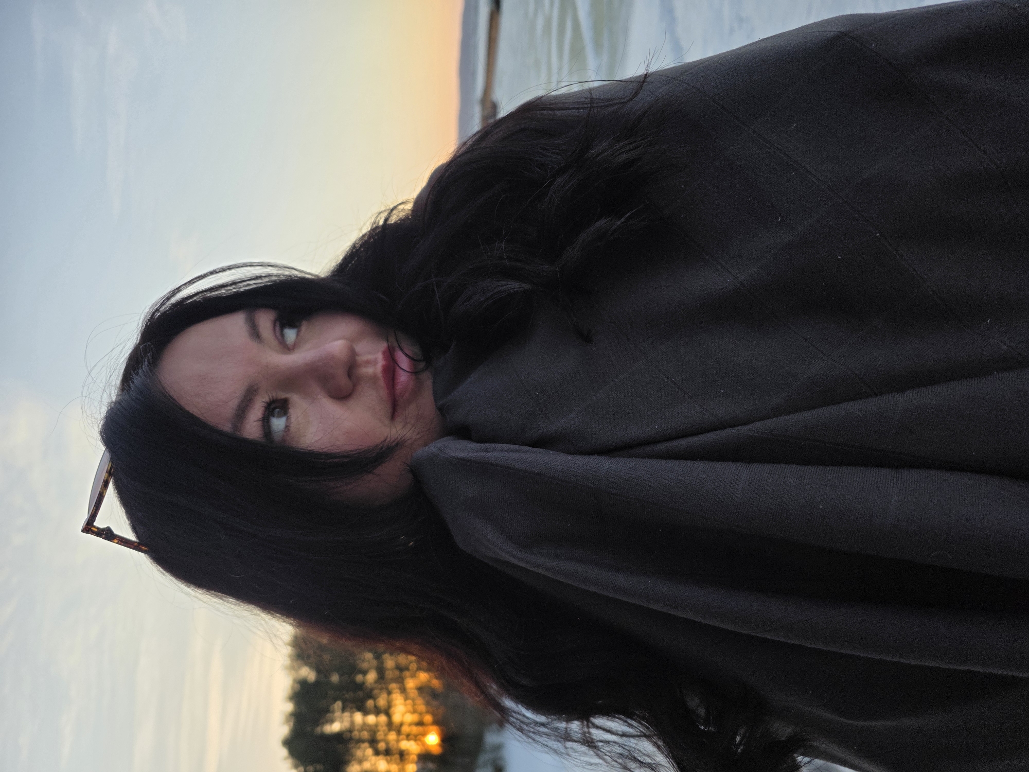 A woman with dark hair, partially covered by a dark fabric, looks to the left in an outdoor setting. Sunglasses are tucked into her hair. The background shows a light-colored ground, possibly snow, with trees and a gradient sky transitioning from blue to orange, suggesting sunrise or sunset. The location is possibly Chaikovsky, Russia.