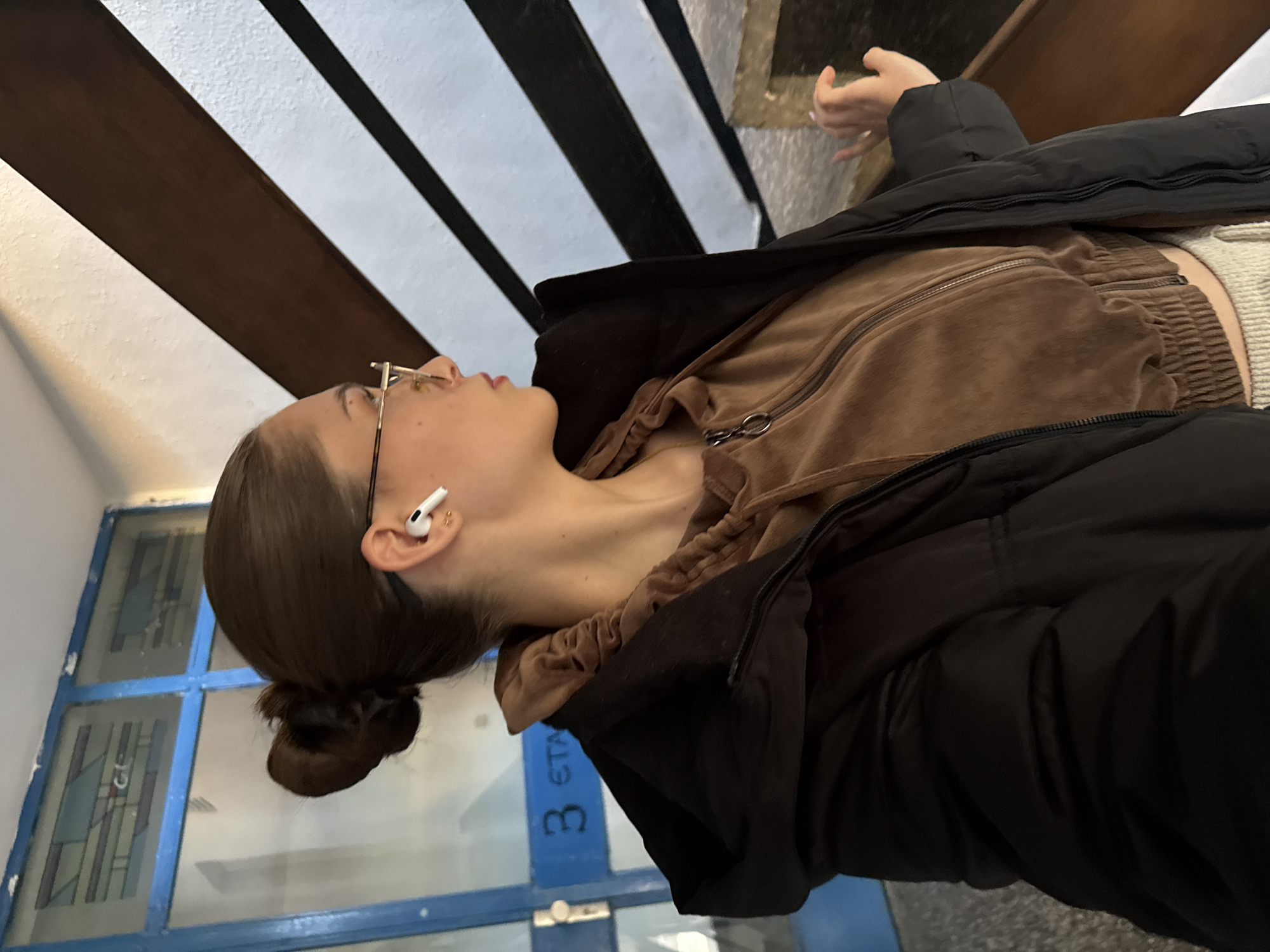 A young woman with light skin, brown hair pulled back in a bun, and wearing glasses and an AirPod in her left ear is lying on her back. She is wearing a brown zippered jacket under a black puffer coat. The woman is lying on a stairwell with blue and white window panes to her left and wood and white steps above her. There is blue text on the wall below her, with a "3" partially visible. The photograph was likely taken in a stairwell within an apartment complex in Sofia, Bulgaria.