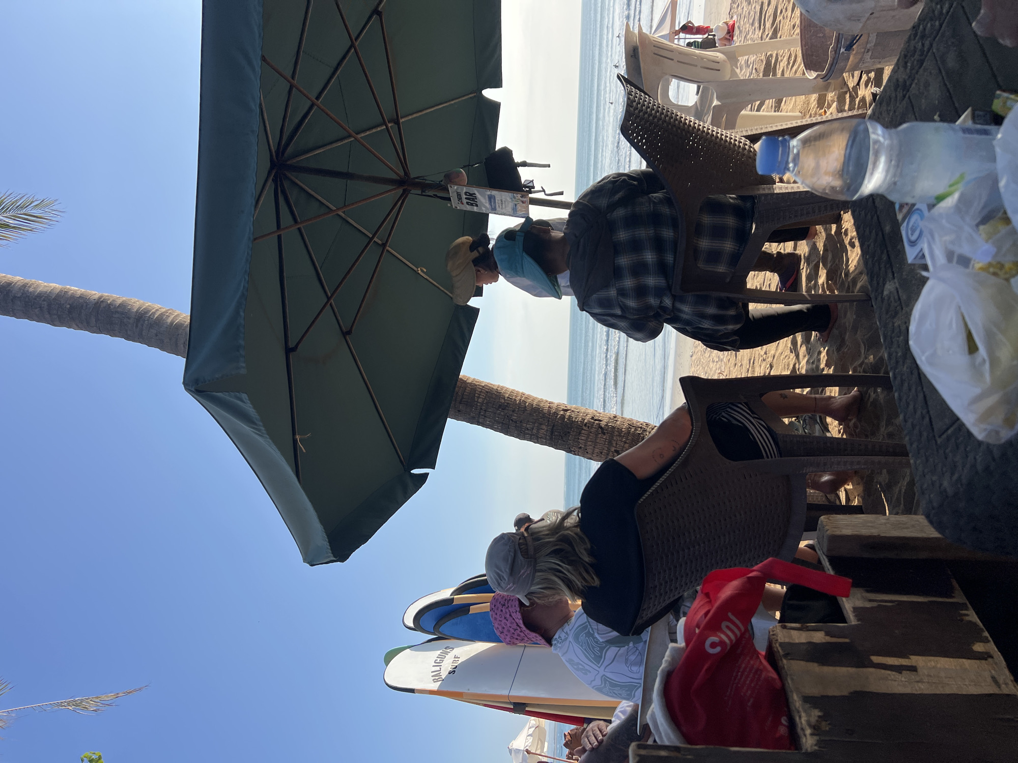 The image depicts a beachfront scene in Kuta, Indonesia, likely on a sunny day given the clear sky and bright lighting. In the foreground, a weathered wooden table is partially visible, laden with plastic bags containing food items and a bottle of water. Behind the table, several people are seated in chairs, facing away from the camera and towards the ocean.
To the left, three surfboards are propped up vertically, one with the text "BALIGUNS SURF" visible. A palm tree trunk stands prominently in the background, leaning slightly. Overlooking the scene is a large, dark green umbrella, providing shade.
Under the umbrella, a woman wearing a hat and a light-colored shirt is standing, possibly serving or interacting with others. Another person in a checkered shirt is seated, facing away. The sandy beach stretches out to the ocean, where gentle waves are rolling in. A few distant figures are visible on the beach and in the water. The overall atmosphere suggests a relaxed, outdoor dining or lounging experience at a beachside establishment.