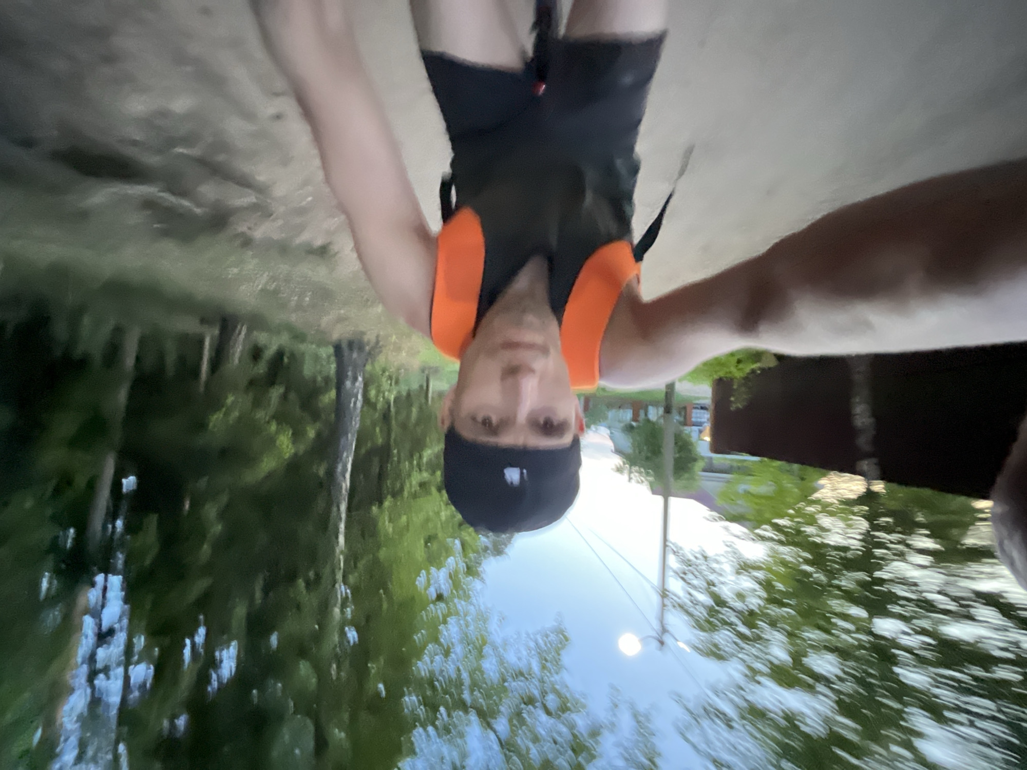 A man wearing a black baseball cap and a black and orange life vest is oriented upside down in the frame. The background appears to be a park-like setting with trees and greenery. A building and a street light are also visible. The location is potentially Chaikovsky, Russia.