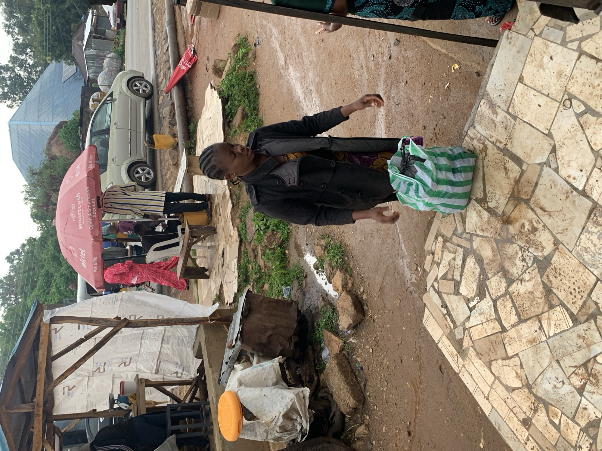 The image depicts an outdoor marketplace scene in Jos, Nigeria. In the foreground, a woman with braided hair, wearing a dark coat over a patterned dress, stands facing left. She holds a green and white striped bag filled with black trash bags. To her left, an orange disc rests on a makeshift stall constructed of wood and white sheeting. Various discarded materials, including corrugated metal and rocks, are scattered on the muddy ground. In the background, a pink umbrella with the "Airtel" logo provides shade over several people and a vehicle. Beyond that, a silver car is parked on a paved road, with more stalls and a large, angular, blue-roofed structure in the distance. The ground appears wet and muddy, suggesting recent rainfall.