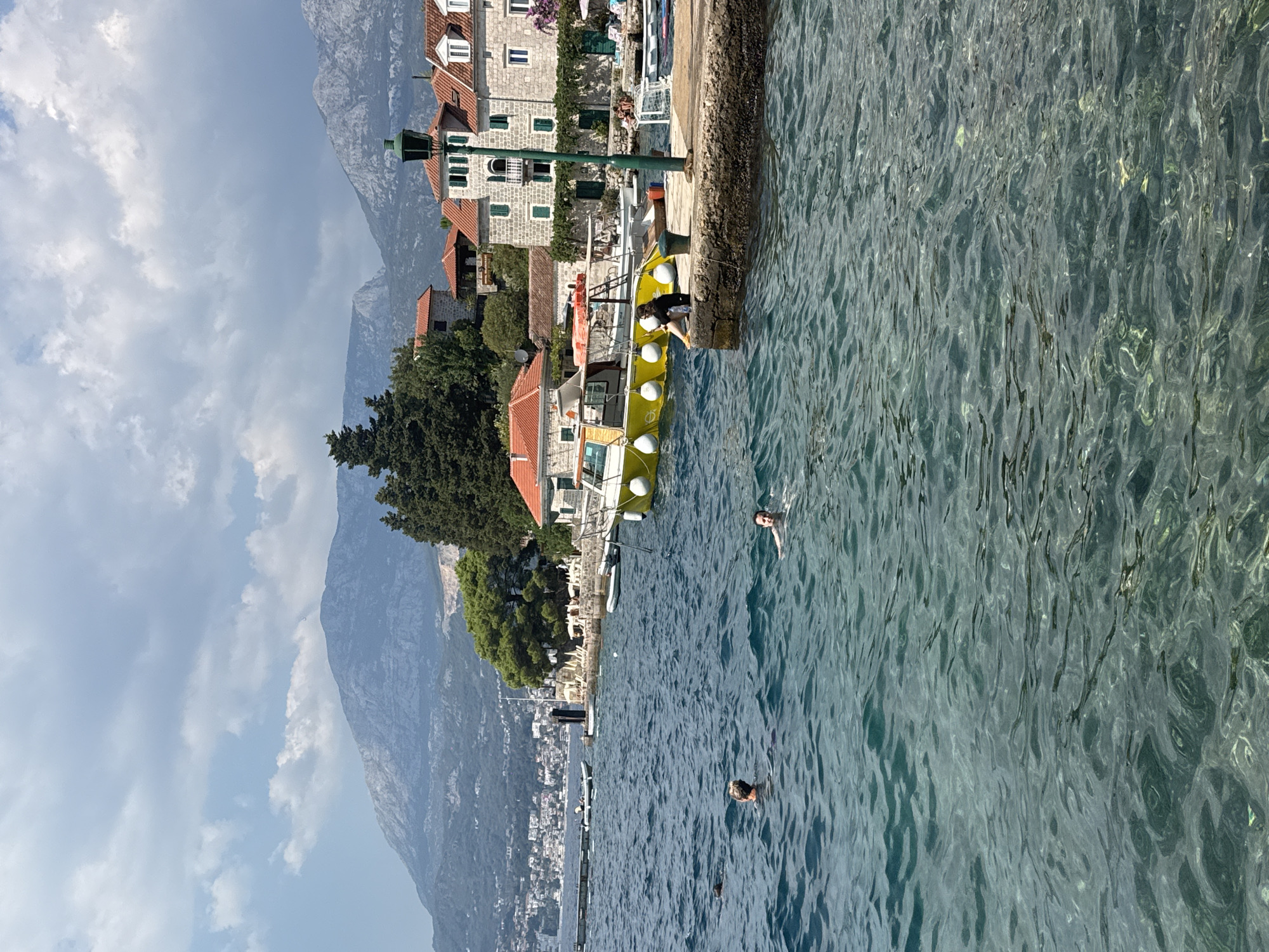 In the foreground, the crystal-clear turquoise water of a bay laps gently. Several people are swimming and enjoying the water. In the middle ground, a yellow boat with white fenders is moored to a stone pier. A person in a black shirt and yellow shoes sits on the edge of the pier, facing the water. Behind the pier, there are several buildings with red-tiled roofs, characteristic of Mediterranean architecture. A tall green lamppost stands prominently on the pier. In the background, a coastal town is visible with more buildings and a dense cluster of green trees. Beyond the town, imposing mountains with rugged slopes rise into a sky filled with dramatic, layered clouds. The scene depicts a sunny day at a scenic coastal location.