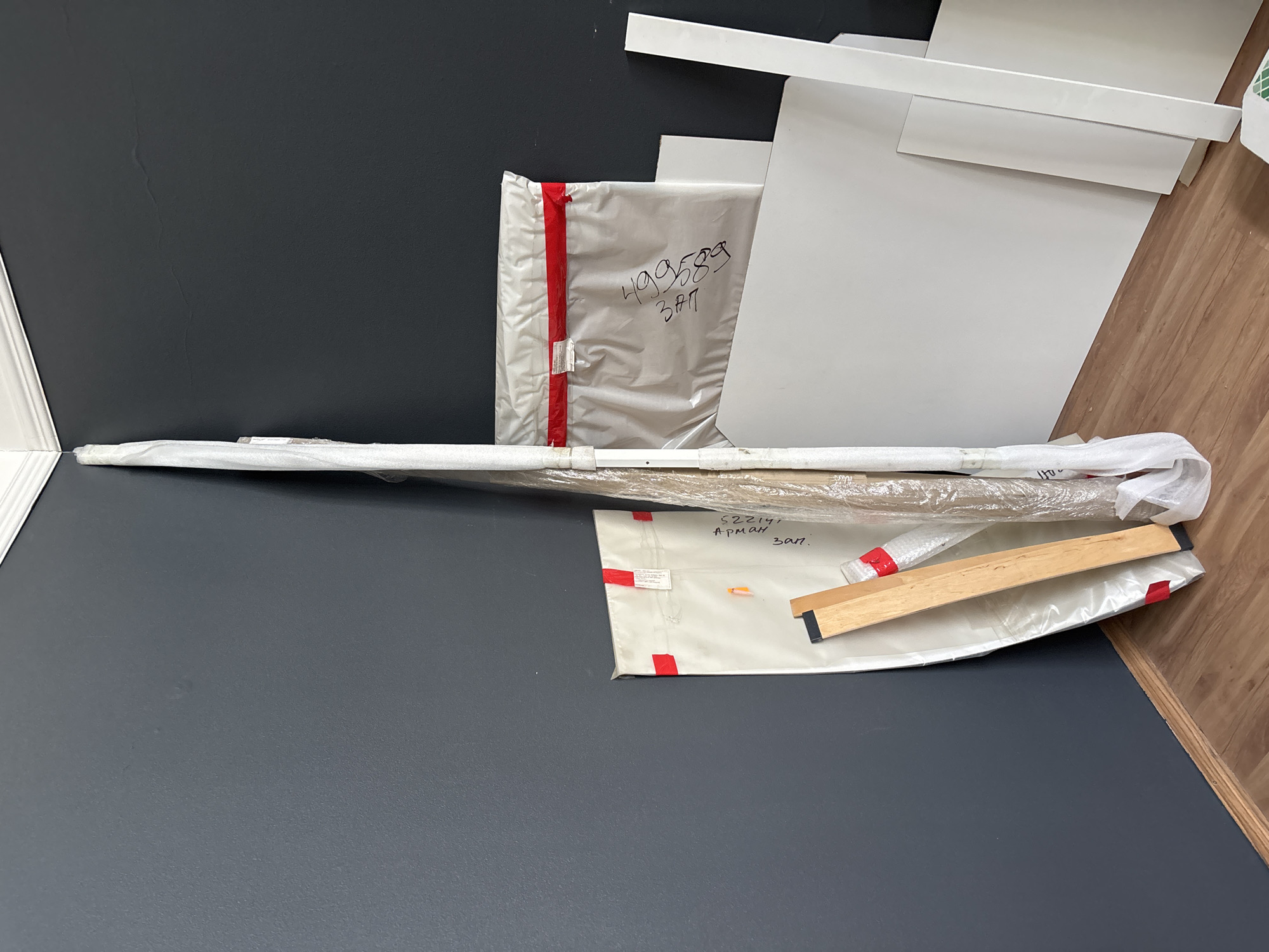 The image shows a corner of a room with dark gray walls and a light brown wooden floor. In the corner, there are several objects leaning against the wall, including long, flat white pieces of material, possibly construction supplies or large artwork. Some of these are wrapped in plastic. There is also a wooden piece with black ends and a long, slender object wrapped in clear plastic. Two white fabric-like bags with red tape are also present; one has black writing that appears to be "499589 3ft" and another has writing that appears to be "Apman 3017". The scene suggests a storage area or a preparation space for a project, likely in an indoor residential or commercial setting. The image does not contain any information to suggest a location in Zaporizhzhia, Ukraine.