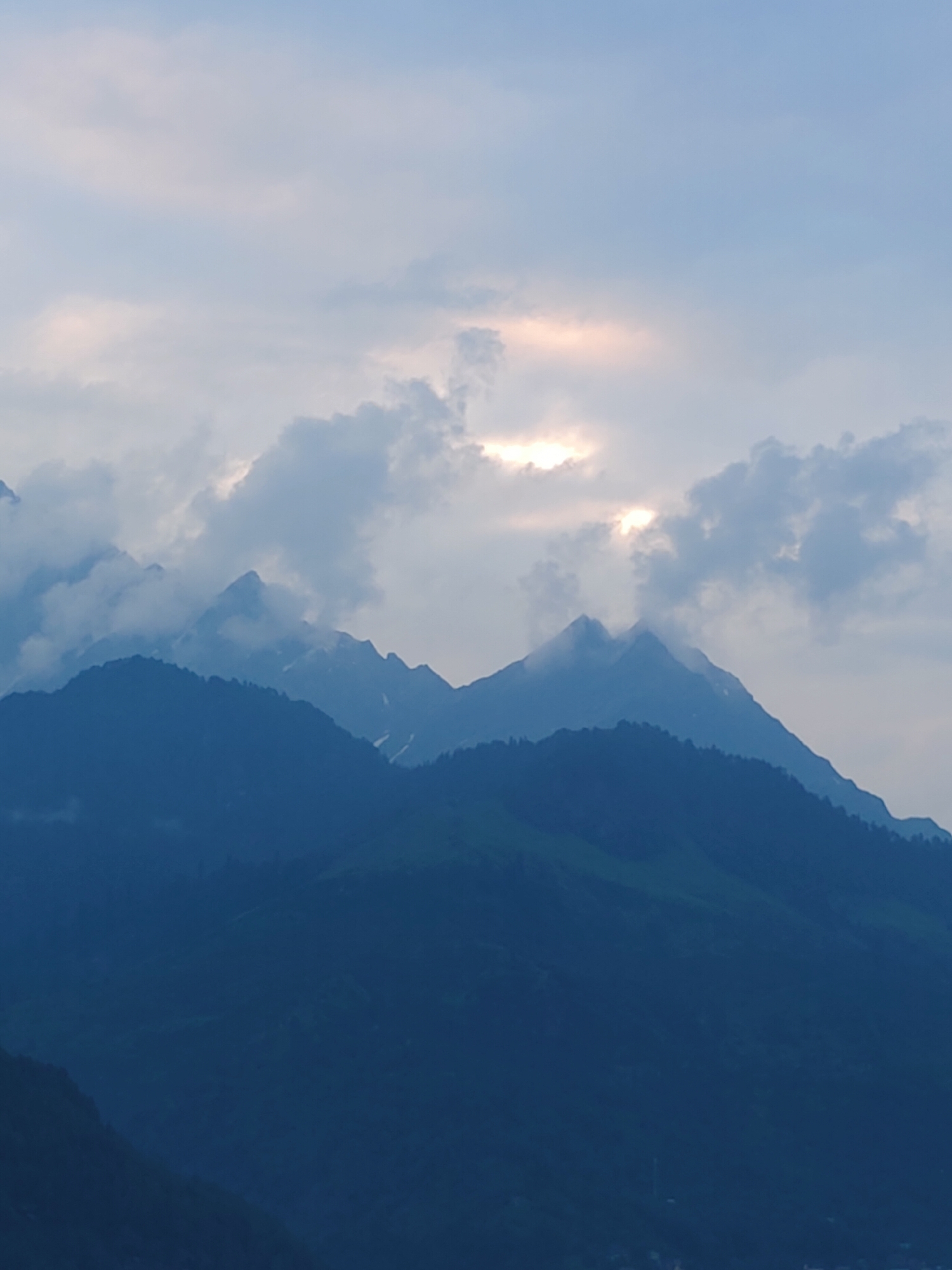 This media file captures a majestic mountain landscape at dusk. The scene is dominated by several layers of mountain ranges, rendered in deep blues and purples, suggesting the fading light. The foreground features the darkest, most solid mountain silhouette, with hints of green vegetation on its slopes. Behind it, progressively lighter layers of mountains rise, some appearing to be shrouded in mist or clouds.
The sky above is a soft, pale blue, with scattered, wispy clouds. A notable detail is a patch of sunlight breaking through the clouds towards the top center of the image, casting a warm, golden glow that illuminates the edges of the clouds and creates a luminous effect. This suggests it's either sunrise or sunset, with the light struggling to pierce through the atmospheric conditions.
There are no people or specific objects visible that would indicate a particular activity. The focus is entirely on the natural grandeur of the mountains and the sky. The overall mood is serene and contemplative, evoked by the dramatic lighting and the vastness of the landscape. The location cues, such as the layered mountains and the quality of light, are consistent with a mountainous region like Vashisht, India, often characterized by such scenic beauty, especially during twilight hours. There is no visible text in the image.