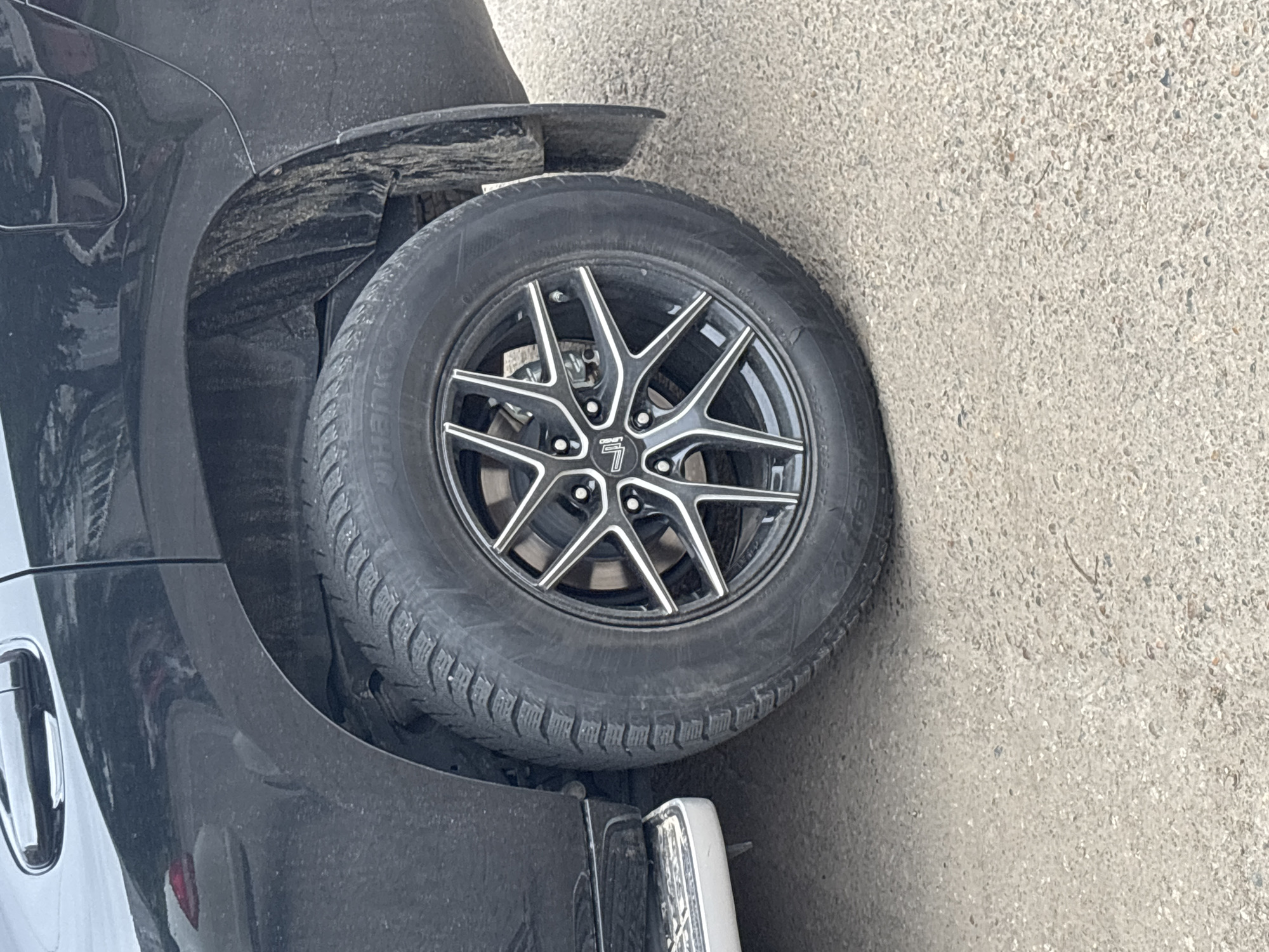 The image shows a close-up of the rear wheel of a black SUV parked on a light-colored, gravelly surface, consistent with a street or parking lot in Blagoveshchensk, Russia. The vehicle's tire is a winter tire with a distinctive tread pattern, and it is fitted with a black alloy wheel with silver accents. The wheel has a multi-spoke design. The vehicle's bodywork, specifically the rear fender and door, is visible, showing a dark, reflective finish. There is some dirt and grime on the car and the wheel well, suggesting recent use in less than pristine conditions. A silver step/running board is partially visible on the lower left side of the image. The environment appears to be outdoors, under daylight conditions, with no other vehicles or people clearly visible in the frame. The overall impression is of a parked vehicle in an urban or semi-urban setting.