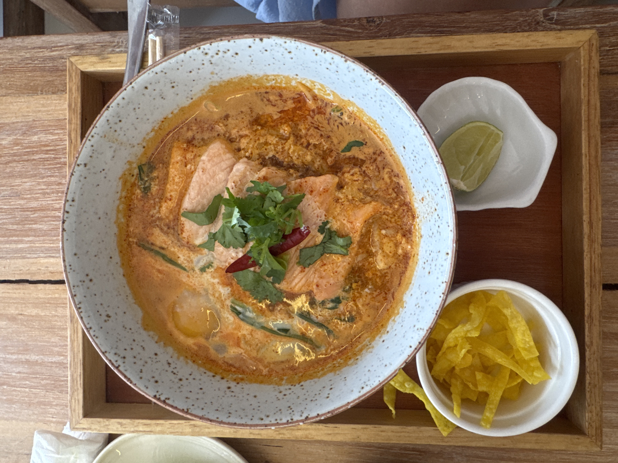 An overhead shot displays a light-colored, speckled bowl of Tom Yum soup filled with broth, salmon pieces, green vegetables, and garnishes of cilantro and dried chilies. The bowl is placed on a wooden tray that sits on a wooden table. To the left of the bowl, a small white dish holds yellow, crispy wonton strips. To the right, another small white dish contains a lime wedge. The scene appears to be in a restaurant setting in Ko Phangan, Thailand.