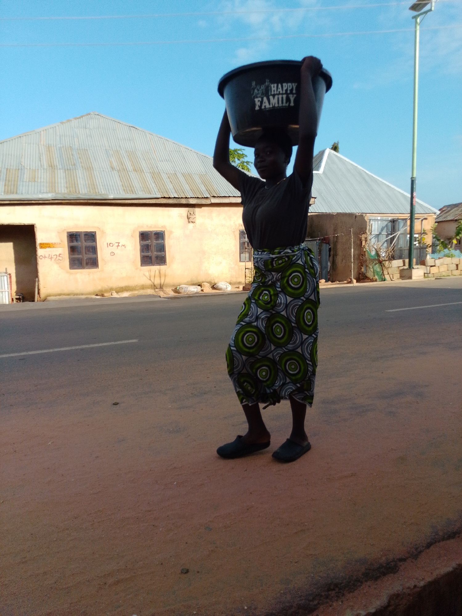 A young Black female, appearing to be a teenager or young adult, is walking on a dirt road, carrying a large black plastic basin filled with water on her head. She is wearing a dark gray t-shirt and a colorful patterned skirt that is green, white, and black. She is also wearing dark slip-on shoes. The basin has the words "HAPPY FAMILY" written in white on its side.
The background shows a row of buildings with corrugated metal roofs, typical of Gombe, Nigeria. One building has two rectangular windows and some writing on its wall. A solar-powered street light stands to the right of the woman. The sky is clear and blue. The scene suggests a daily activity in a residential area, likely in the city of Gombe.