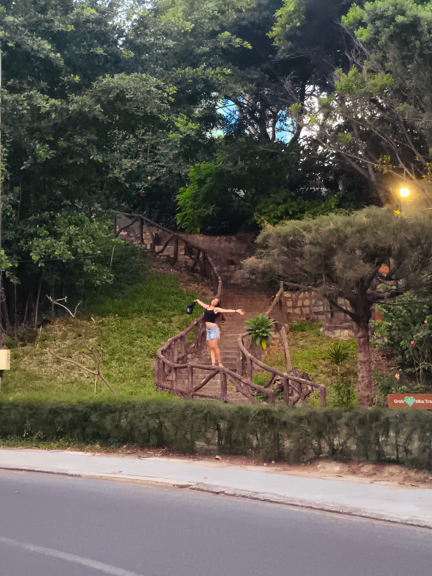 A young woman stands on a wooden staircase that winds up a grassy, tree-covered hill. She is wearing a black crop top and denim shorts, with her arms outstretched to her sides. She appears to be smiling, and her hair is dark and shoulder-length. The staircase has wooden railings and is bordered by lush green vegetation. In the background, there are tall trees, stone walls, and glimpses of the sky. The scene is brightly lit, suggesting it is daytime. A street is visible in the foreground, with a hedge separating it from the hill. The location is identified as Bac Nha Trang Ward, Vietnam.