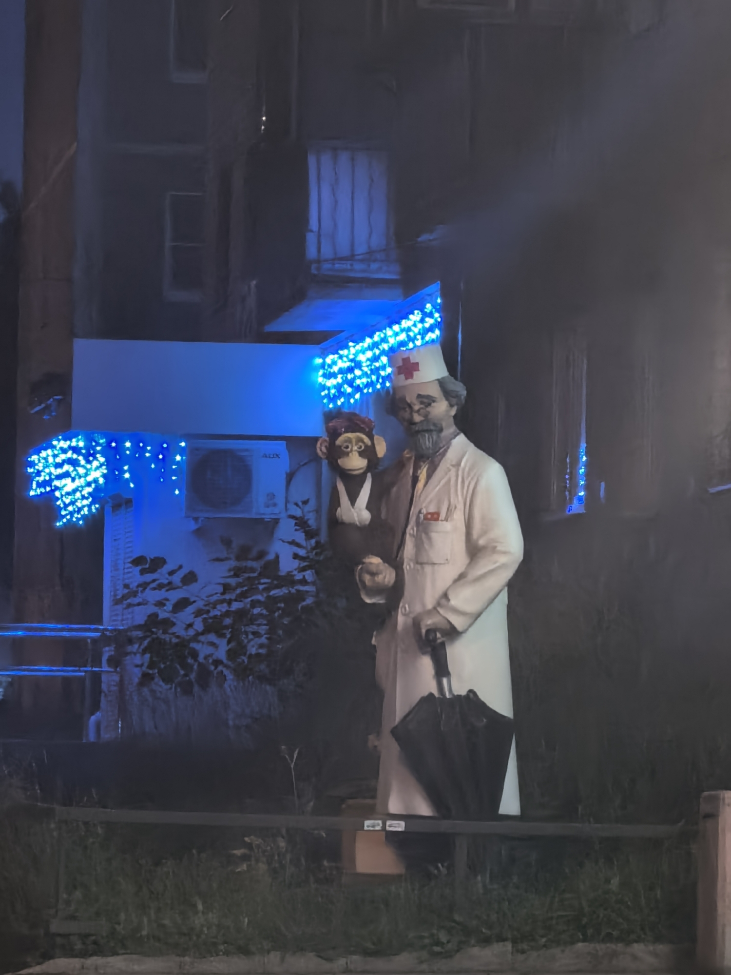 This is a full-shot, eye-level, outdoor photograph of a statue of a man dressed as a doctor and a statue of a monkey. The doctor statue is on the right, wearing a white coat, a cap with a red cross, glasses, and a beard. He is holding a black umbrella in his right hand and a small object in his left hand. The monkey statue is on the left, wearing a hat and what appears to be a white smock. The statues are positioned in front of a building that has blue decorative lights strung across its exterior. An air conditioning unit is visible on the wall behind the statues. The ground in front of the statues is covered in grass, and there is a dark metal railing. The overall lighting is dim, with the blue lights providing a significant source of illumination. The scene appears to be at night or in low-light conditions.