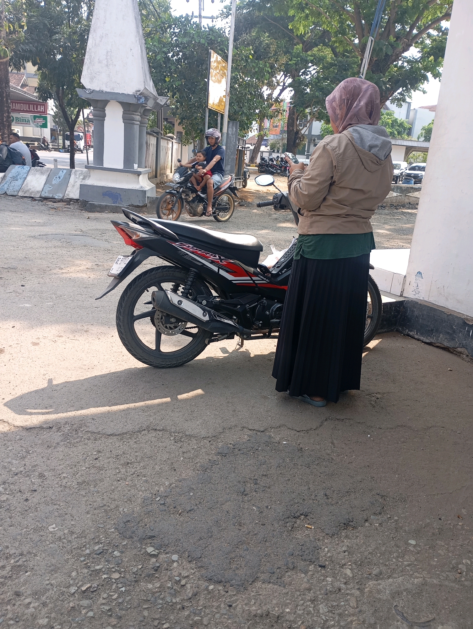 The image captures a street scene in Pacitan, Indonesia, under a bright, sunny sky. In the foreground, a woman wearing a hijab, a tan jacket over a green shirt, and a long black skirt stands next to a black and red motorcycle. She is looking down at a smartphone in her hands. The motorcycle, positioned at an angle, has its rear wheel and exhaust pipe clearly visible.
In the mid-ground, another motorcycle is being ridden by a man wearing a blue shirt and a helmet, with a young child sitting in front of him. They are on a paved road, with trees and buildings lining the background. To the left, a white and grey concrete monument with decorative pillars stands prominently, with a sign displaying "HAMDULLILLAH" and "BIMA" visible on a building behind it. Other parked motorcycles are also present.
The ground is a mix of rough asphalt and concrete, with some debris scattered around. Sunlight casts shadows on the pavement, indicating the time of day. The overall impression is a snapshot of everyday life on a street in a town or city in Indonesia.