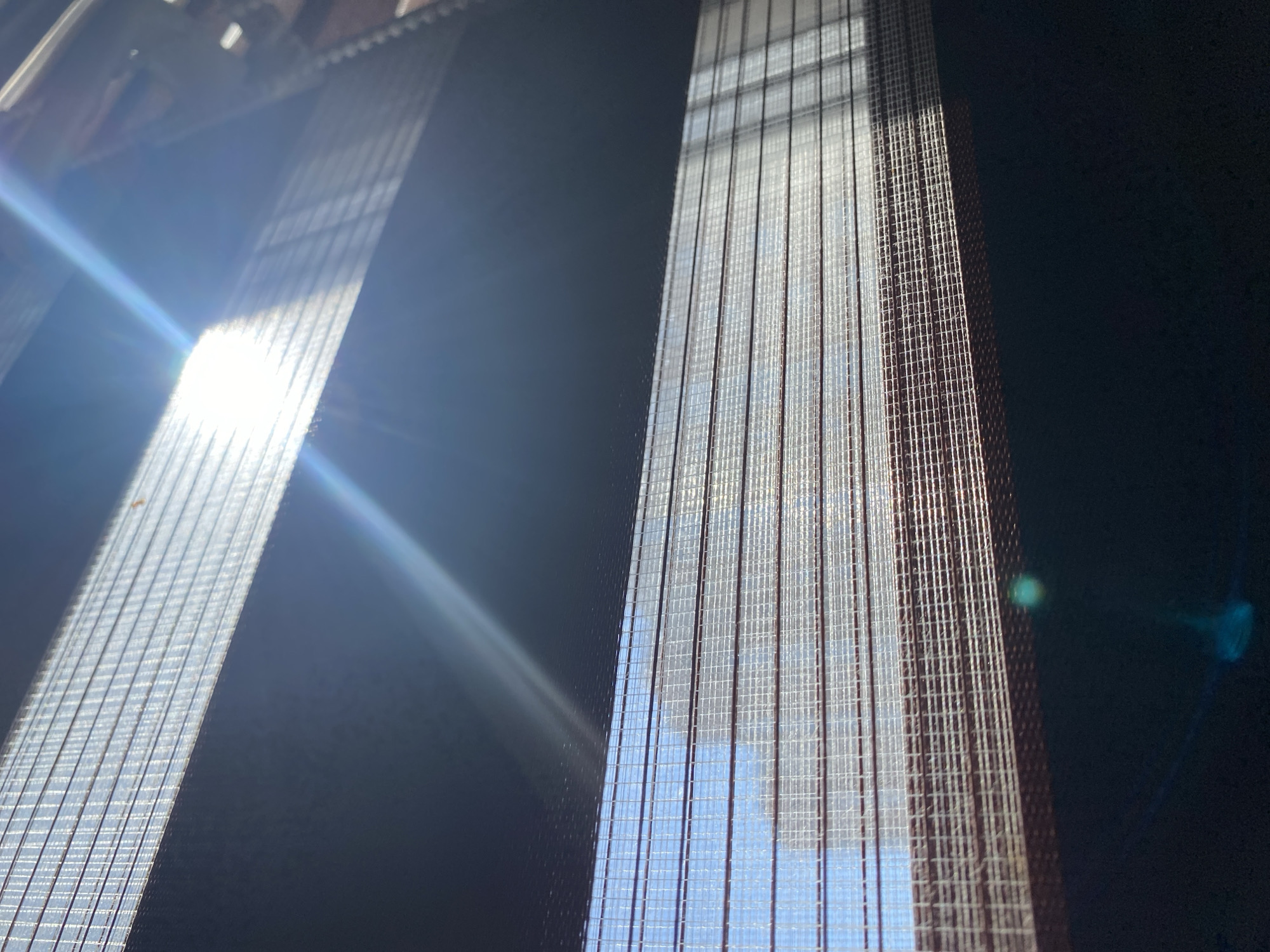 The image shows two partially illuminated vertical blinds against a dark background. Sunlight streams through the blinds on the left, creating a bright glare and emphasizing the grid-like structure of the fabric. The blinds on the right are more evenly lit, allowing a clearer view of the woven texture and the sky visible through them. The background is dark, possibly the interior of a room. No people, actions, or interactions are visible. The provided information does not offer enough detail to confirm if the image was taken in Chaikovsky, Russia. The focus is on the light and texture of the blinds.