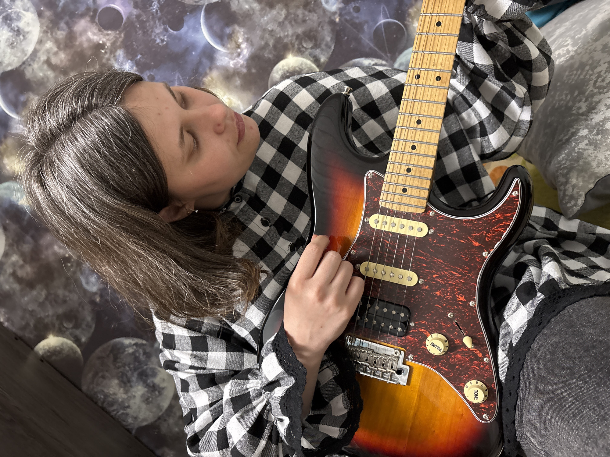 A young woman with graying brown hair, wearing a black and white checkered flannel shirt with lace trim, is lying down and holding an electric guitar. Her eyes are closed. The guitar is a sunburst Stratocaster style with a maple neck and a tortoiseshell pickguard. The background appears to be a wall decorated with an image of celestial bodies. Based on the prompt, this scene takes place in Chaikovsky, Russia.